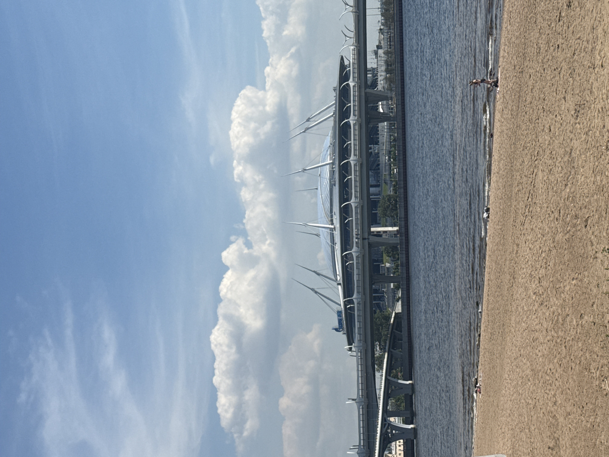 The image shows a wide shot of the Gazprom Arena stadium in Saint Petersburg, Russia, on a sunny day with a partly cloudy sky. The stadium, with its distinctive saucer-like roof and pointed supporting structures, is situated across a body of water, likely the Neva River. A multi-level bridge complex spans the water in front of the stadium. In the foreground, there is a sandy beach with a few people visible. One person stands near the water's edge, while others are seated or lying on the sand. The water is rippled, indicating a slight breeze. The overall scene captures a modern urban landscape with recreational activity.