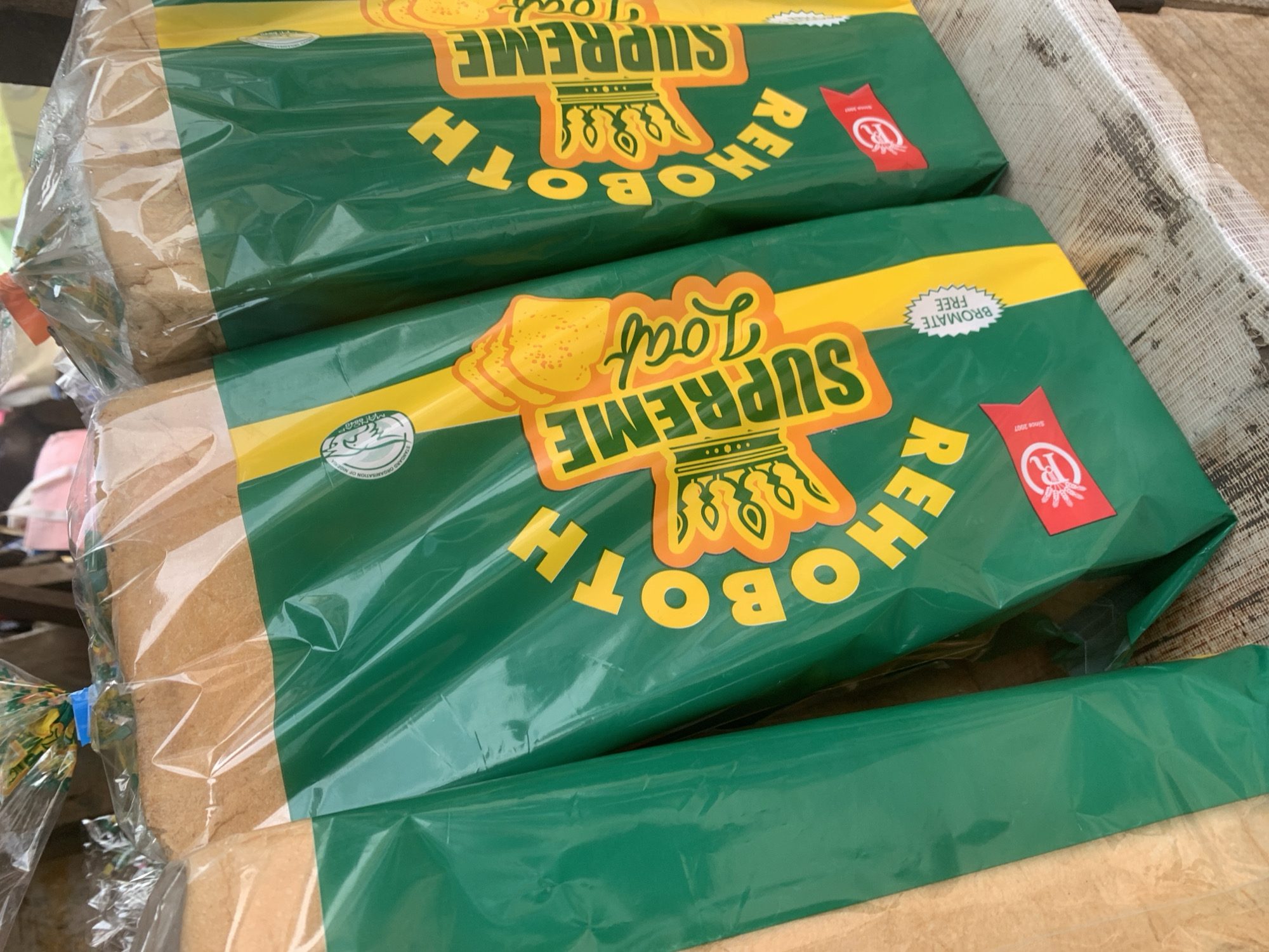 The image displays three loaves of Rehoboth Supreme Loaf bread, packaged in green and yellow plastic bags. The bread bags feature prominent yellow lettering spelling out "REHOBOTH SUPREME LOAF" and illustrations of bread slices and a crown. The loaves are arranged side-by-side, with the leftmost loaf partially visible. A white, serrated sticker on the middle loaf reads "BROMATE FREE". Red ribbons with a circular logo are present on the lower portion of the middle and rightmost loaves. The bread is positioned on a wooden surface, likely a market stall. The background is blurred, showing hints of other market items and possibly people. The overall scene suggests a retail environment in Shagamu, Nigeria, where these loaves of bread are being sold.