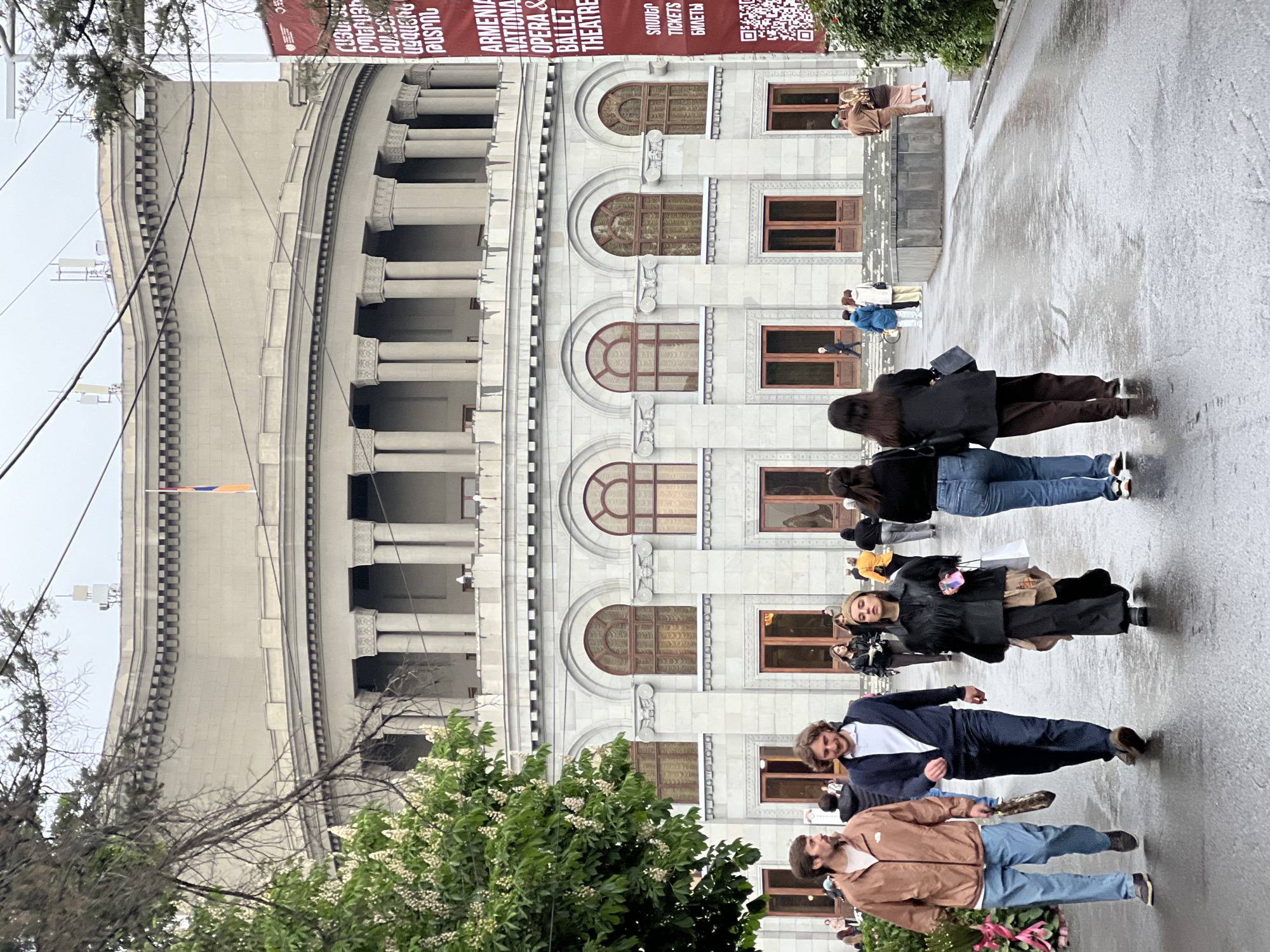 A medium, outdoor shot shows pedestrians walking on a street in front of a large, grey building in Yerevan, Armenia. The building is the Armenian National Opera & Ballet Theater. The architectural style is neoclassical with rounded arches over the windows and columns along the top of the building. A banner hangs on the side of the building promoting opera and ballet performances. There is also a scannable QR code. A small, thin orange, blue and white flag is visible. On the bottom left of the image, some greenery, trees with flowers, can be seen. The street is wet. There are seven visible pedestrians. Two male pedestrians are in the foreground, with the man on the left wearing a brown jacket and jeans and holding flowers, and the man on the right wearing a blue suit. Behind them are five more pedestrians, with the first person wearing black. In the distance, more pedestrians are visible in front of the building.