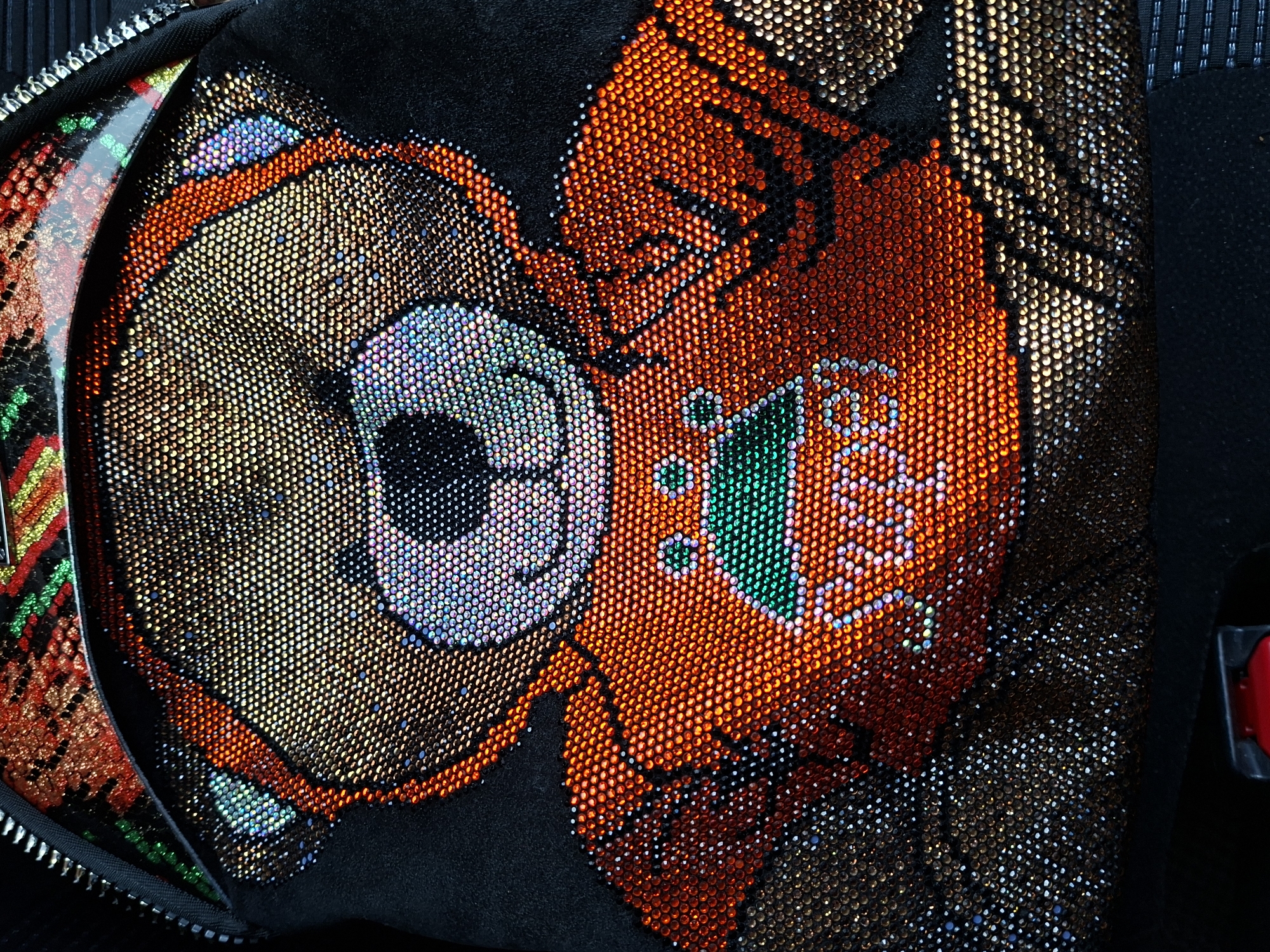 The image is a close-up, high-angle shot of a black backpack adorned with rhinestones. The focal point is a design of a teddy bear's face and upper body. The teddy bear's face is outlined in silver and light blue rhinestones, with black rhinestones for the eyes and nose. The ears and body are covered in shimmering gold and orange rhinestones. The bear is wearing an orange shirt with a green crown emblem on the chest, and the word "Super" is written in white rhinestones below the crown. The top of the backpack, visible at the top of the image, features a colorful, striped pattern with red, orange, green, and yellow rhinestones. A silver zipper runs along the top left edge of the backpack. The background is dark and out of focus, but appears to be a dark surface, possibly the interior of a car or furniture, with a small section of a red object with a black strap visible at the bottom center. There is no indication of the city of Barnaul, Russia, or any specific location within it.