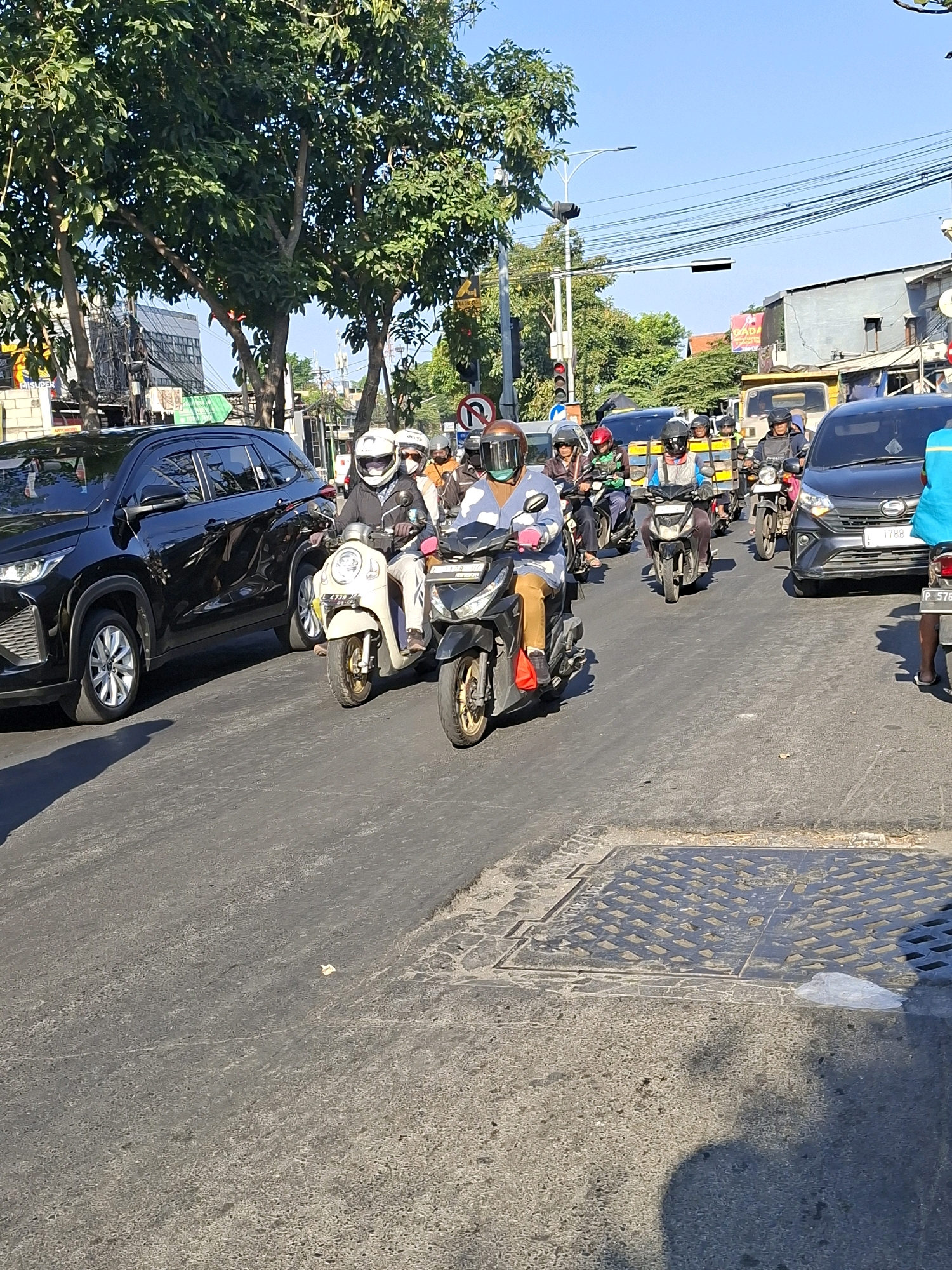The image depicts a street scene in Surabaya, Indonesia, likely during daylight hours. Several motorcycles and a black SUV are visible on a paved road, suggesting traffic congestion. The motorcycles are carrying riders, most of whom are wearing helmets and some appear to be wearing masks. One motorcycle in the foreground has a light-colored body and a rider with a white helmet. To the left, a black SUV is parked or in slow-moving traffic. In the background, there are trees, buildings, power lines, and traffic signals, indicating an urban environment. The road surface shows signs of wear and tear, with some manhole covers present. The sky is clear and blue. The overall impression is of a busy street in a city.