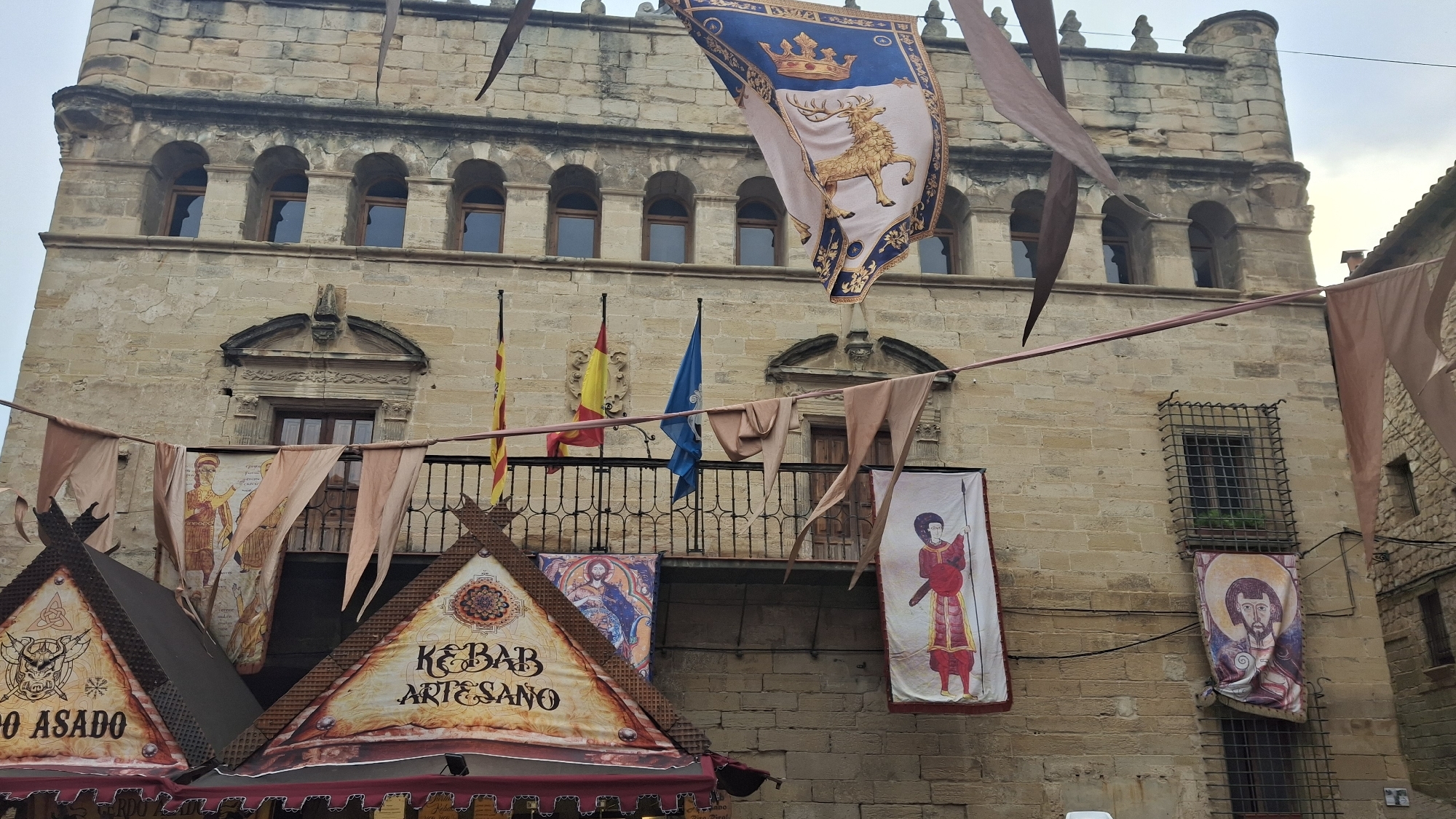 In the city of La Fresneda, Spain, a traditional stone building serves as the backdrop for a festive scene. Banners and flags adorn the facade, creating a medieval or historical ambiance. Prominently displayed is a large blue and gold banner featuring a crowned stag, likely a heraldic symbol. Several smaller flags, including a yellow and red one, flutter in the breeze. Fabric streamers in muted tones are strung across the foreground, adding to the decorative atmosphere. Below, a market stall or awning with triangular roofs is visible, adorned with colorful banners depicting figures and text, including "Kebab Artesano." The building itself has arched windows and decorative architectural details, suggesting it is a historic structure. The sky is overcast, indicating potentially mild weather.