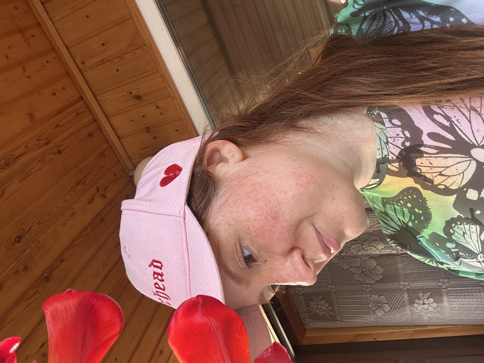 A woman with reddish-brown hair is wearing a pink baseball cap with a red heart on the side and the word "Ahead" embroidered in red. She is wearing a green and lavender butterfly-patterned shirt. The background consists of a wooden ceiling and window, along with several artificial red tulips. It is not possible to determine the location of the picture with the given information.