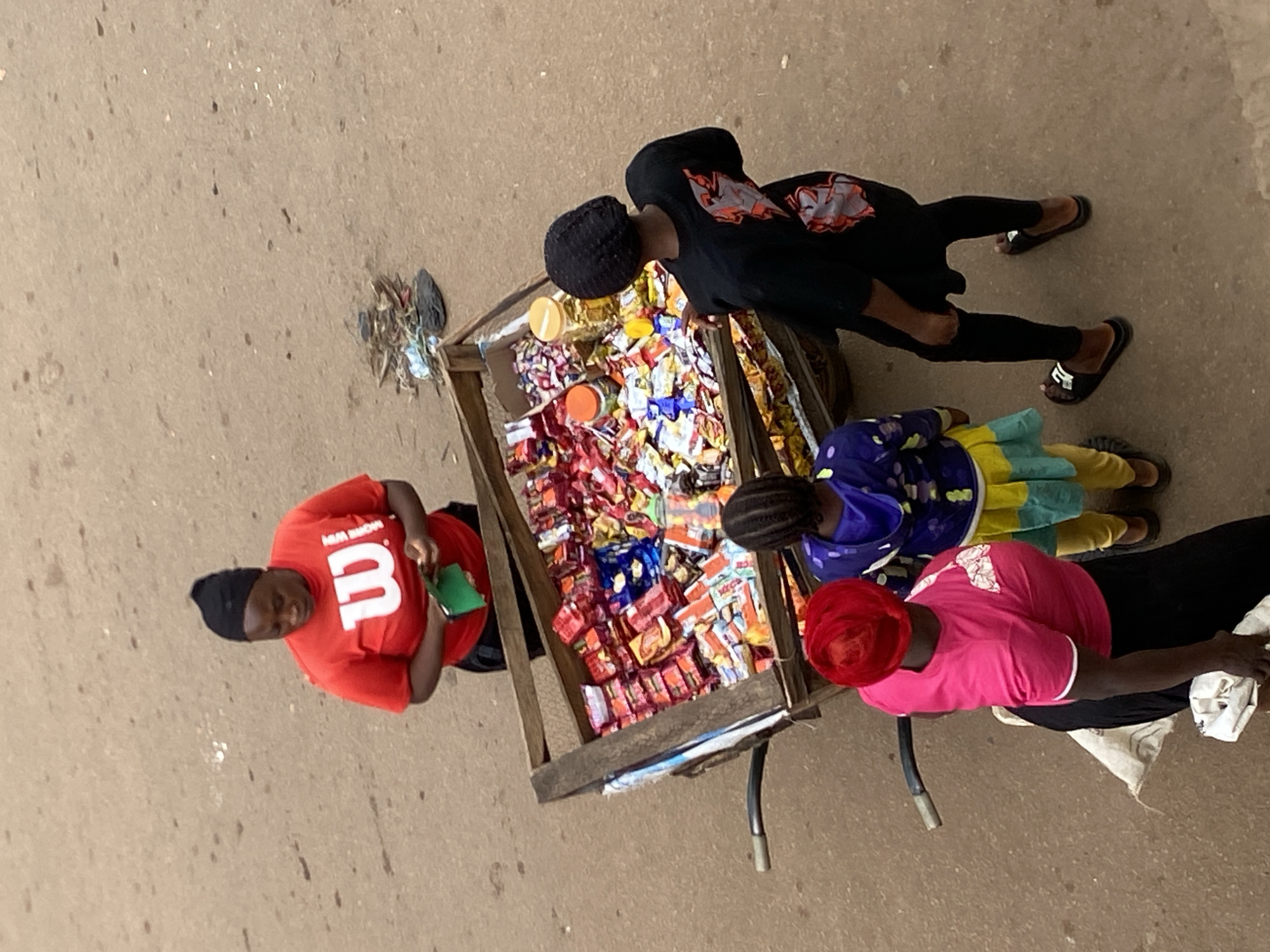 In Bukuru, Nigeria, a vendor stands by a wooden cart brimming with an assortment of packaged snacks. She wears a red t-shirt with a white "W" logo and a black headscarf, holding a green wallet or pouch. In front of the cart, two children and a woman observe the merchandise. The woman wears a bright pink t-shirt and a red headscarf. One child, facing away, is dressed in a blue and yellow outfit. Another child, also facing away, wears a black t-shirt with a graphic design and black leggings, and black slides. The ground is a sandy, uneven surface, with scattered debris and what appears to be a discarded plastic bag and some loose paper. The cart has metal handles and appears to be on wheels, suggesting it's a mobile stall. The scene is captured from an elevated, top-down perspective.