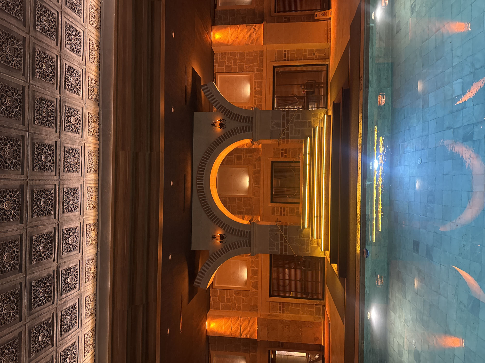 The image shows a building exterior in Ürgüp, Turkey. The building features a decorative facade with carved stone panels on the left and a stone structure with arches in the center. The arches are illuminated with warm light. To the right is a swimming pool reflecting the building's lights. The scene is captured at night, and the lighting creates a warm, inviting atmosphere.
