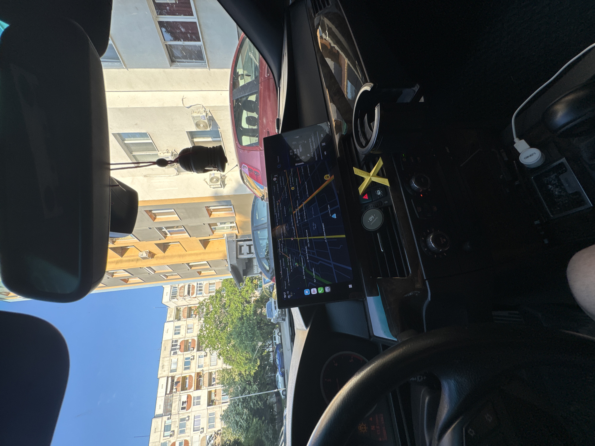 The image depicts the interior of a vehicle, specifically from the driver's perspective, looking outward through the windshield towards an urban street scene on a clear, sunny day.
Within the vehicle's interior, a modern, large aftermarket infotainment screen is centrally mounted on the dashboard. The screen displays a navigation map, clearly indicating streets in Cyrillillic script, including "бул. Патриарх Евтимий" and "ул. Св. Княз Борис I", confirming the location as Stara Zagora, Bulgaria. Other visible street names include "ул. Радецки", "ул. Ген. Гурко", "ул. Сливница", "бул. Цар Симеон Велики", "ул. Христо Ботев", and "ул. Васил Априлов". The time displayed on the screen is 19:17. Below the map, a music player interface is visible, showing "PREEK" and "Непознато". A yellow "X"-shaped decorative object is placed on the dashboard near the central air vents, and a black cupholder is positioned to its right. A dark, decorative air freshener hangs from the rearview mirror. A white USB-C charging cable with a "LOND" brand adapter is plugged into a port in the center console area, lying on a dark carpeted floor. The steering wheel is partially visible on the left side of the frame, showing a portion of the instrument cluster. Only a knee/leg of an individual is visible in the bottom right corner, indicating the presence of a person in the driver's seat.
Outside the vehicle, a street is lined with multi-story buildings. To the left, a large, light-colored residential building with numerous balconies and external air conditioning units is prominent. To the right, a building with a yellow-orange facade and white windows, also equipped with air conditioning units, is visible. Several vehicles are parked on the street, including a red Fiat Punto directly in front of the yellow building, a silver SUV further down the street, and a blue sedan. Green trees with dense foliage are interspersed between the buildings and along the street. The sky above is clear and blue.