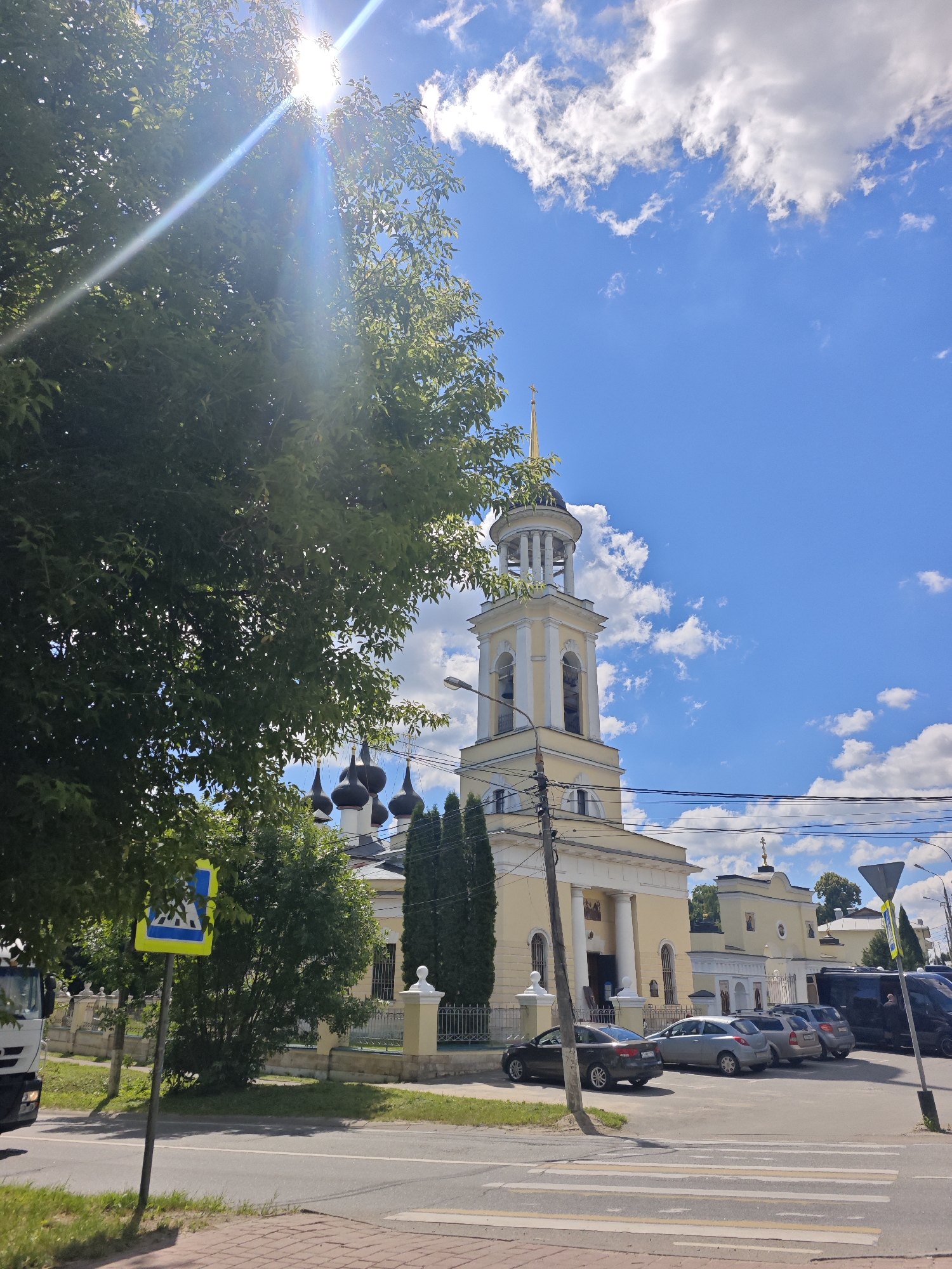 In Chekhov, Russia, under a bright blue sky dotted with fluffy white clouds, a yellow church with a tall bell tower stands prominently. The sun's rays streak through the leaves of a large, dark green tree in the foreground, partially obscuring the view of the church. A pedestrian crossing sign, a blue and yellow diamond shape with a depiction of people crossing, is visible on the left side of the image. Several cars are parked in front of the church, and a white truck is partially visible on the far left. The street in front of the church has a marked pedestrian crossing. The church has multiple dark domes and a golden cross atop the bell tower. Electrical wires and a lamppost are also visible near the church.
