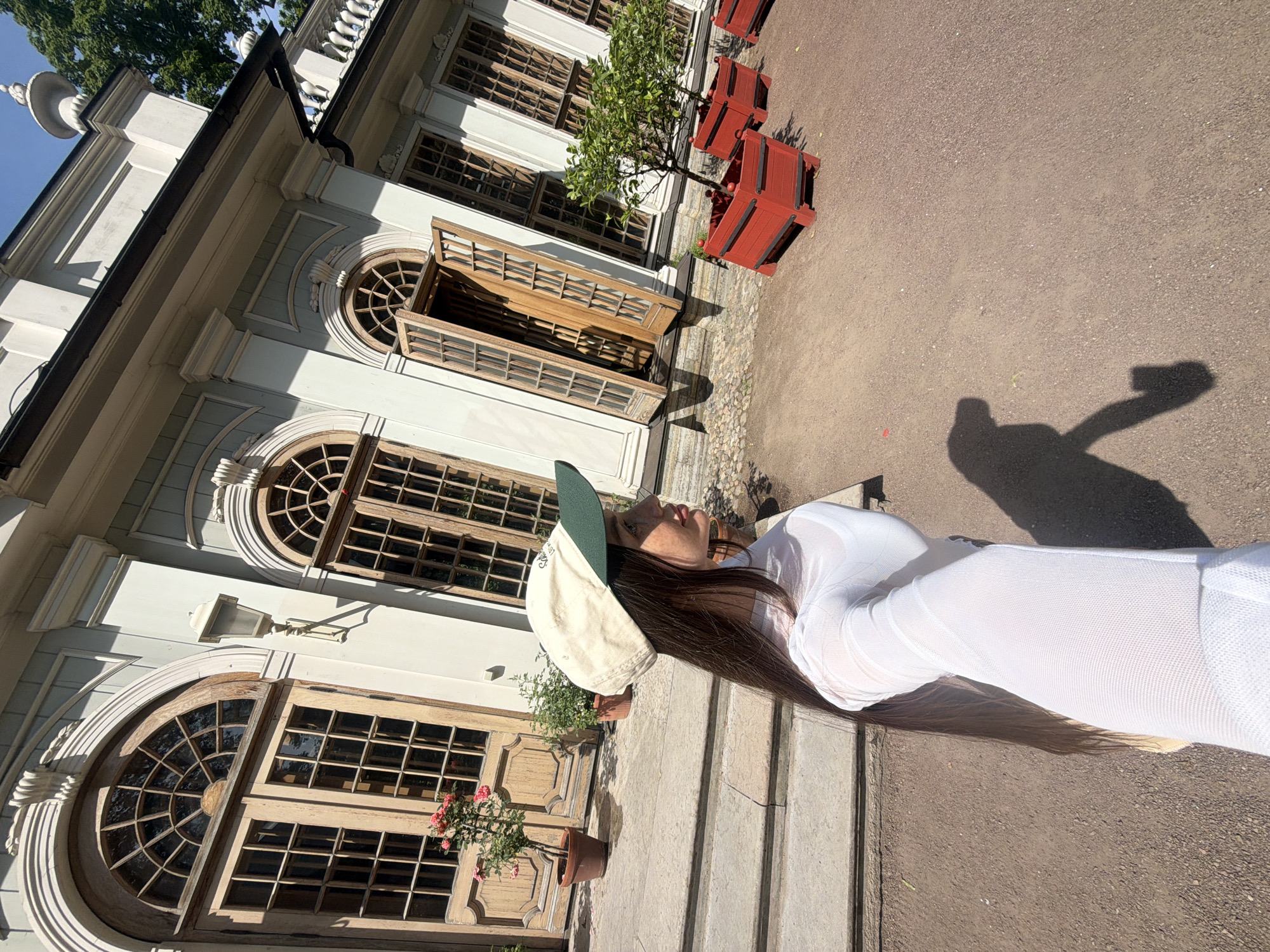 In Saint Petersburg, Russia, a young woman with long, dark hair and a white long-sleeved mesh top is taking a selfie. She is wearing a light-colored baseball cap with a dark green brim. The woman is standing on a gravel path in front of a light-colored building with ornate architectural details, including arched windows with wooden frames and a decorative balustrade. Two potted plants, one with pink flowers, sit on the steps leading up to the building. Several red planters containing small trees are also visible on the path. The scene is bathed in sunlight, casting shadows on the ground.