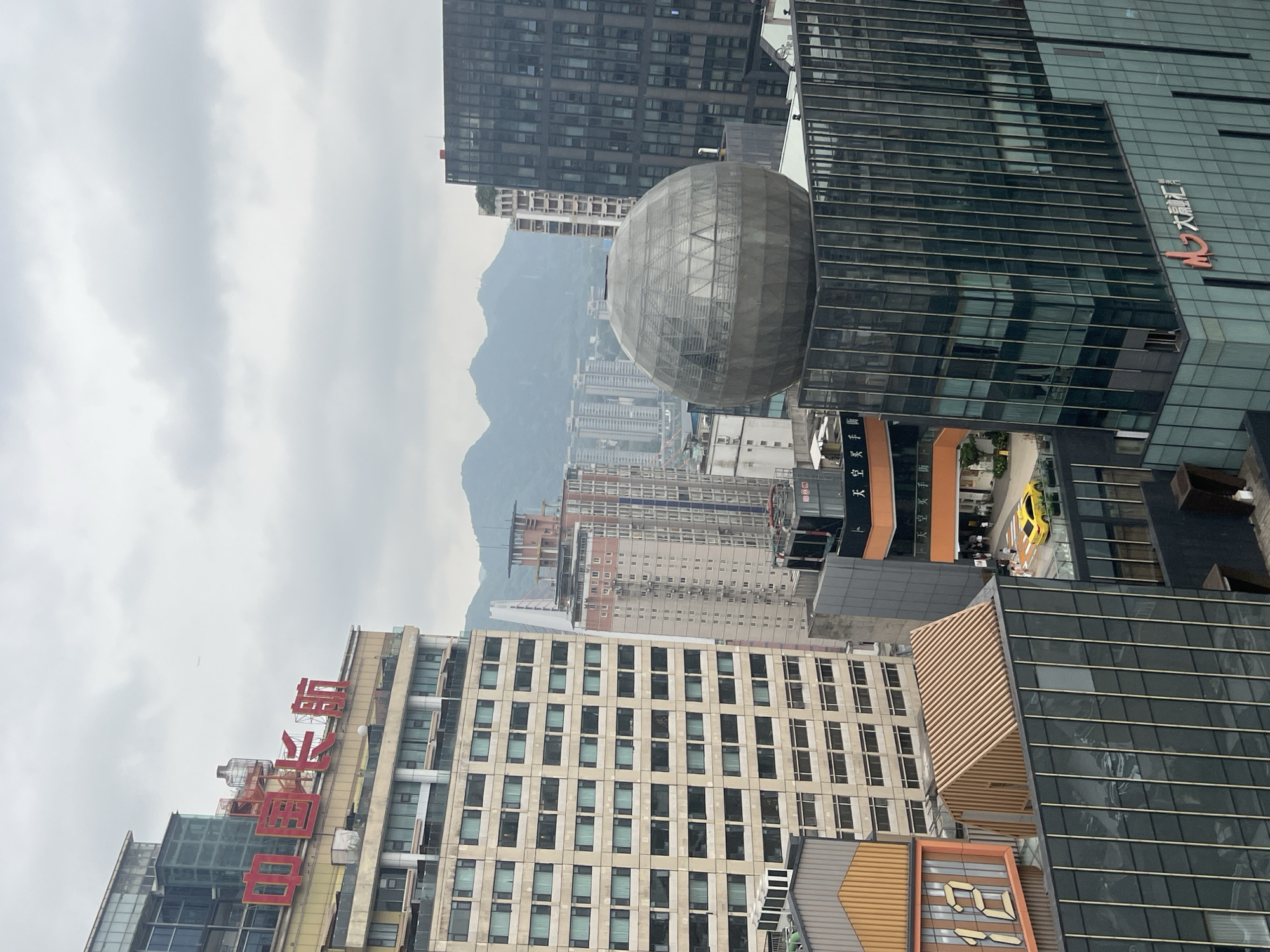 The image depicts a cityscape with several multi-story buildings and distant mountains under a cloudy sky. In the foreground, a large, modern building with a glass facade dominates the right side of the frame. It features vertical metallic accents and a prominent red Chinese logo. Above this building, a large, spherical structure made of intersecting metal supports and glass panels is visible. To the left, a taller, older building with a beige exterior and rows of windows stands out. Red Chinese characters are prominently displayed on its upper section. Further to the left, another building displays a large orange sign with the number "12" in yellow. A small wooden-roofed structure is also visible on top of a building. In the middle ground, several other buildings of varying heights and architectural styles can be seen, including one with a pink and gray facade. A yellow car is parked on a street level between buildings in the mid-ground. The distant background shows a range of hazy, green mountains. The scene appears to be an urban environment in Yuzhong District, China.