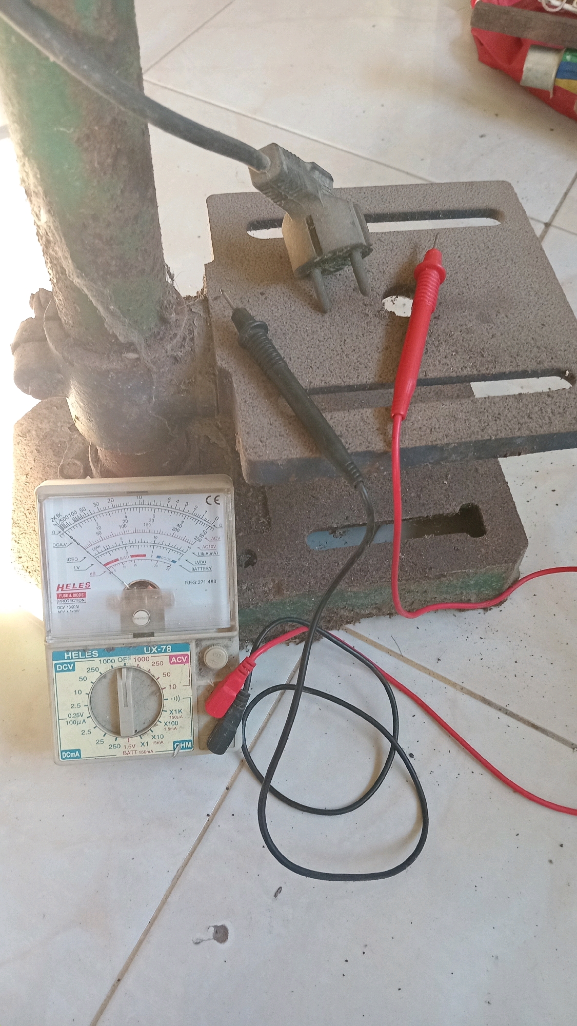 An analog multimeter with a black and red probe is placed on a dusty tiled floor, next to a green metal structure and a rough, square-shaped object. The multimeter, identified as a HELLES UX-78, displays a needle pointing towards a low value on the DCV scale. A grey electrical plug with two prongs is positioned on the square object. The environment appears to be outdoors or in an unkempt indoor space, indicated by the dust and the tiling. The lighting suggests it's daytime. No individuals are present in the scene. The location is identified as Randuagung, Indonesia.