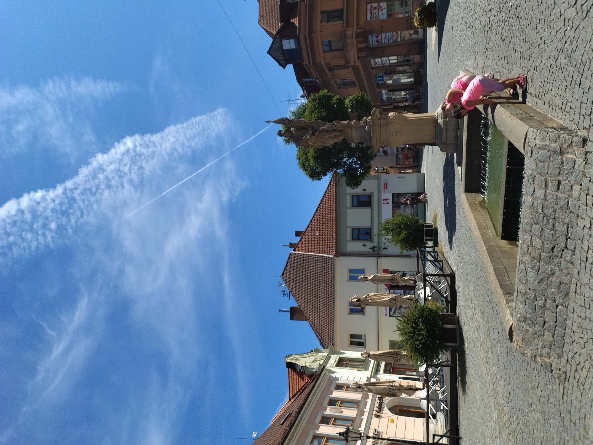 The image depicts a sunny day in the city of Písek, Czechia. In the foreground, two young girls wearing pink tops and shorts are bending over a stone fountain, seemingly playing with the water. The fountain is rectangular with a rough stone border and the water is clear with ripples. The ground is paved with cobblestones. In the middle ground, there is a statue on a pedestal, surrounded by trees. The background features several buildings with terracotta tiled roofs, some of which appear to be shops or restaurants, with outdoor seating areas covered by beige umbrellas. A contrail is visible in the bright blue sky, streaking across a formation of white, wispy clouds. The overall scene is one of a public square on a warm, clear day.