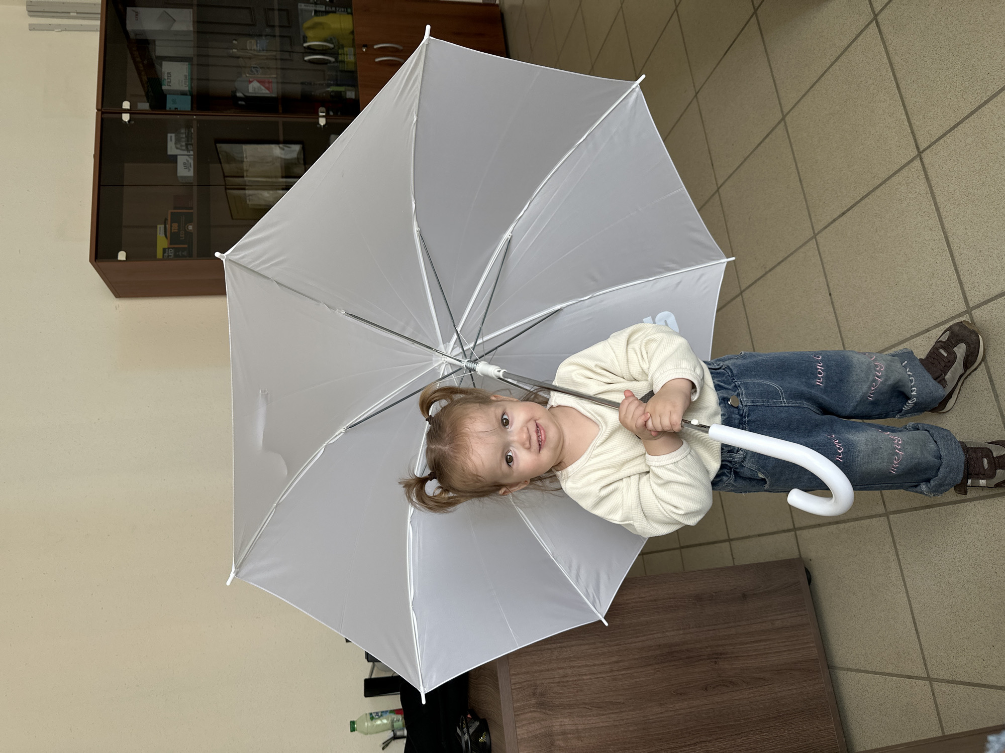 A young Caucasian girl with blonde hair tied in two pigtails smiles at the camera while holding an open white umbrella. She is wearing a cream-colored waffle-knit shirt and blue denim jeans with pink embroidered text. She is standing indoors, in front of a wooden desk and a tall, brown glass-fronted cabinet filled with various items. The floor is tiled with a beige and brown grid pattern. The overall lighting suggests an indoor, well-lit environment, possibly an office or storage area in Yekaterinburg, Russia.