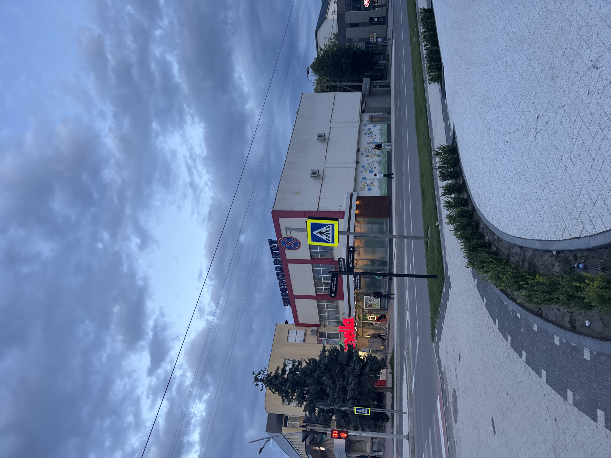 A medium shot captures a street scene at dusk, with a moody sky filled with dark, streaky clouds. The perspective is from a slightly elevated position, looking down a street lined with buildings and a wide, curved sidewalk.
The primary subject is a supermarket with a red and white facade. Above the entrance, the word "SUPERMARKET" is written in large blue letters. There's a pedestrian crossing sign with a yellow triangle and a white silhouette of a person walking. A traffic light is visible, showing red.
On the sidewalk to the right of the supermarket, there are several people. One person appears to be walking a dog. The sidewalk is paved with light-colored stones in a geometric pattern and bordered by neatly trimmed green bushes.
Further down the street, other buildings are visible, some with signs. One sign is partially obscured, but the word "VILNIUS ENTRANCE" is legible on a smaller signpost. Red neon lights are visible in the window of a shop.
The overall mood is one of transition, as day turns into night. The overcast sky suggests impending or recent rain, adding to the somber atmosphere. The streetlights are not yet fully illuminated, hinting at the early stages of evening.