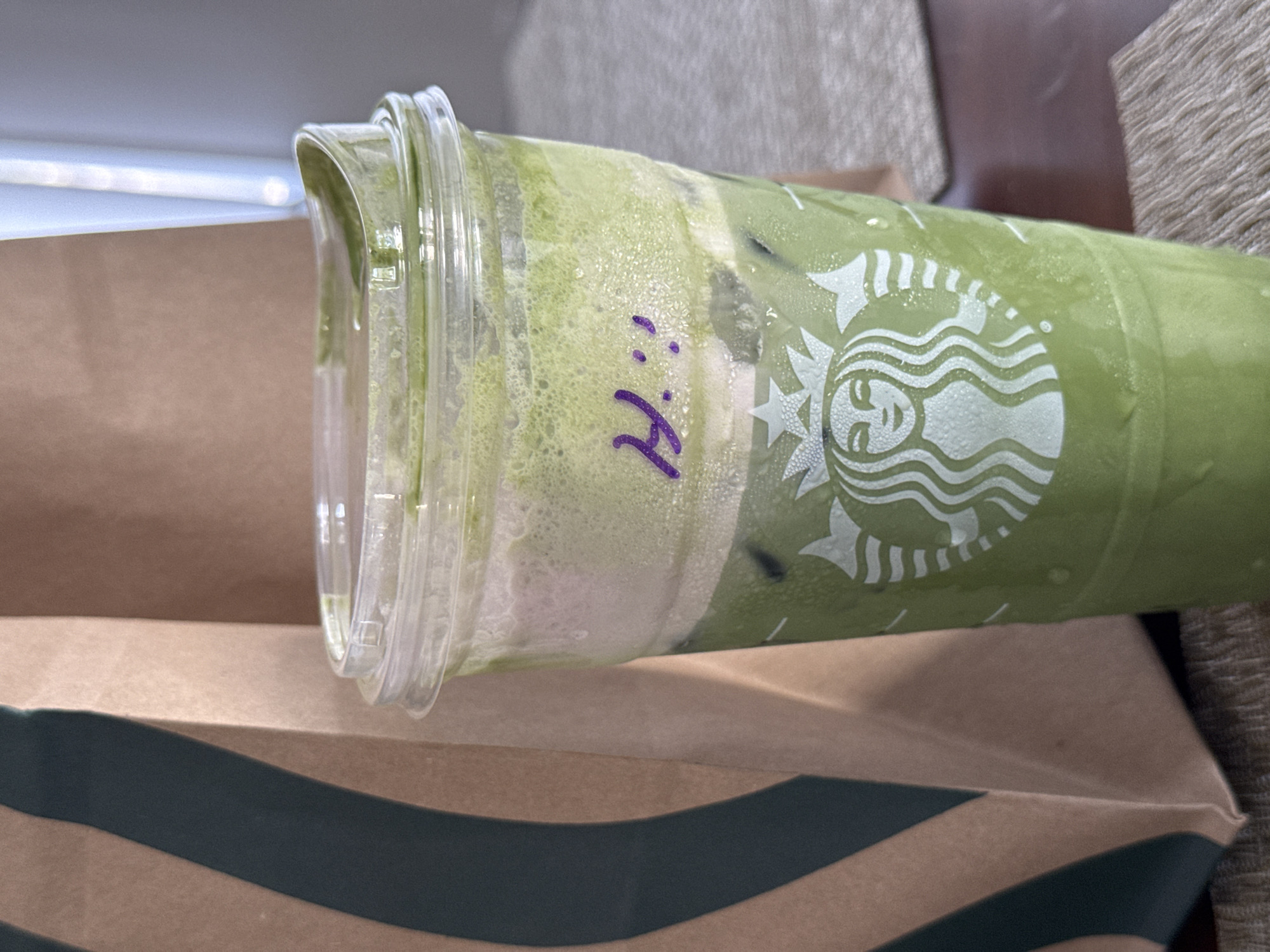 A slightly tilted view captures a Starbucks cup on top of a Starbucks bag in what appears to be an indoor setting. The cup is filled with a green drink that appears frothy with condensation on the cup. The Starbucks logo is visible. "Hi" is written on the cup in purple marker. The bag is brown with a dark green stripe. The setting is likely somewhere in Chula Vista, United States of America.