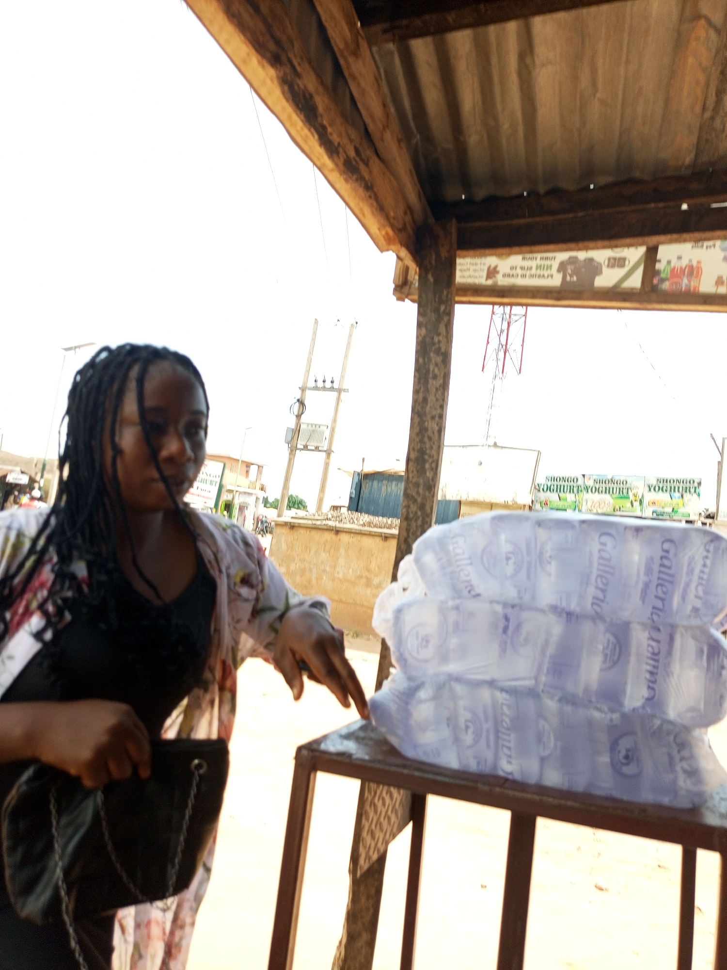 A young woman with dark braided hair stands under a makeshift wooden and corrugated metal shelter. She is wearing a black top and a floral patterned open-fronted shirt. She holds a black purse with a chain strap in her left hand, and her right hand is extended towards a stack of bottled water. The bottles are clear plastic with blue labels. In the background, there are signs for "SHONGO YOGHURT", electrical poles, and buildings, suggesting an urban or semi-urban setting. The ground appears to be sandy or dirt. The overall scene depicts a market or roadside stall environment, possibly in Gombe, Nigeria, given the context provided, with the woman interacting with a vendor or presenting goods.