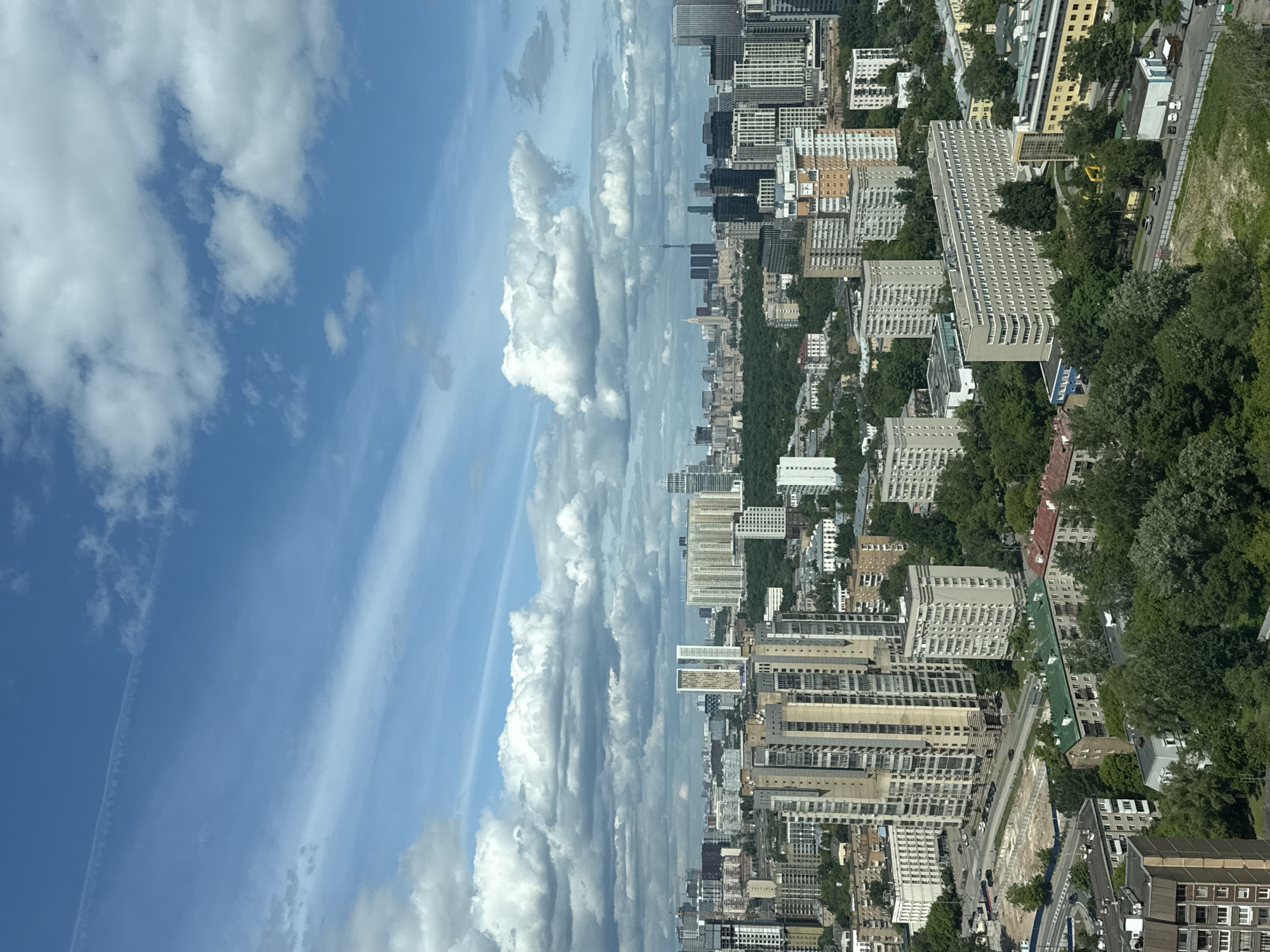 The image presents an aerial view of a cityscape juxtaposed with a cloudy sky, the boundary between the two bisecting the frame vertically. On the right side of the image is the city, characterized by a dense arrangement of buildings, including apartment blocks and other structures, amidst green areas interspersed with trees. Roads and vehicles are visible among the buildings, indicating urban activity. The left side of the image is dominated by a blue sky filled with numerous cumulus clouds. The image is rotated 90 degrees counterclockwise. The location is likely Khimki, Russia.