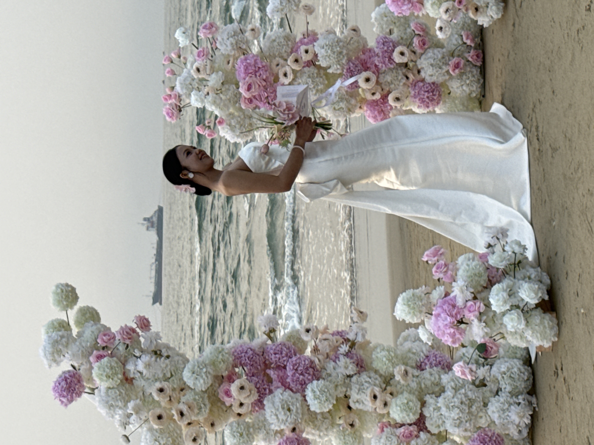 The image shows a woman in a white dress holding flowers. She is positioned in front of a floral arch on a beach. The ocean and a ship are visible in the background. The beach is sandy, and the sky is overcast. The floral arch is composed of white and pink flowers. The scene is at a beach in Đà Nẵng, Vietnam.