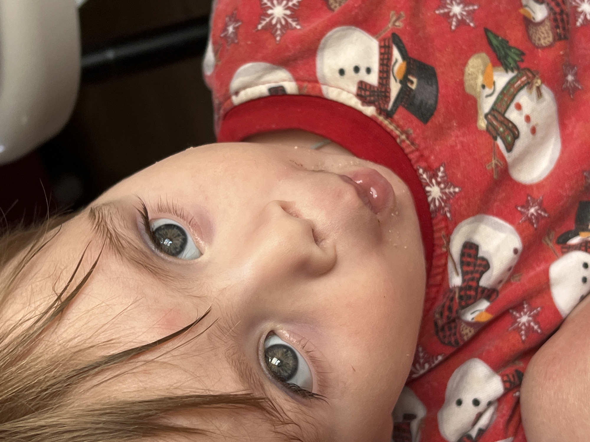 A close-up, slightly tilted portrait captures a young child, likely a baby or toddler, in an indoor setting in Noviy, Russia. The child, the primary subject, has light brown hair and striking blue-grey eyes, wide open and looking directly at the camera with a neutral, inquisitive expression.
The child is wearing a red pajama top or shirt patterned with festive winter and holiday motifs. These include white snowflakes and various characters: snowmen with red plaid scarves and black top hats, and some figures resembling penguins in snowman-like attire. Clear moisture, possibly saliva, is visible on the child's lower lip and chin, along with small white specks around the mouth and nose, which could be dried food remnants or powder.
The background is mostly out of focus, revealing a light-colored object partially visible in the upper left and a dark area behind the child. The setting is clearly indoors, and the clothing suggests a colder season or holiday period. There is no discernible activity other than the child's focused gaze, implying a moment of observation or interaction with the photographer. No text is visible in the image.