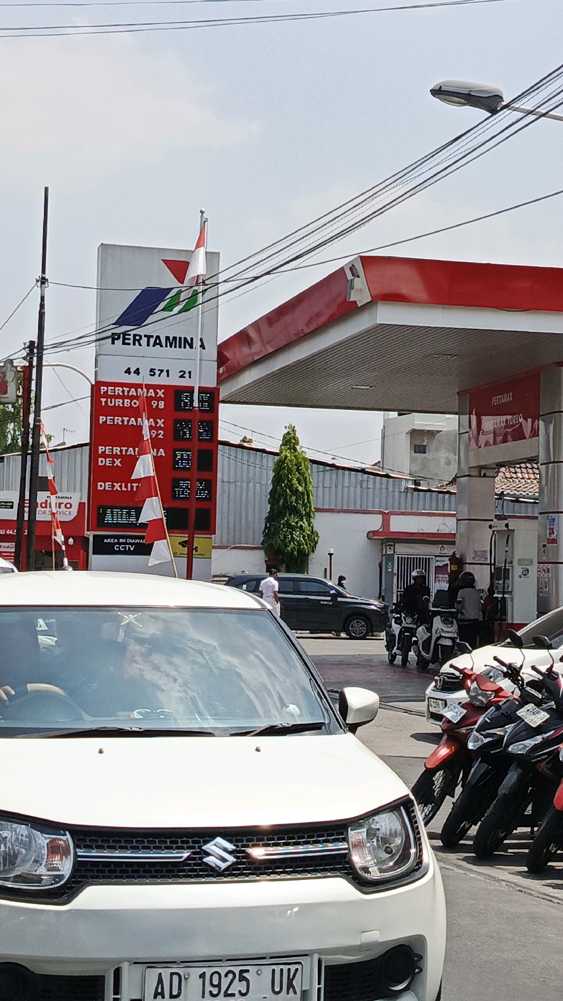 The image shows a busy Pertamina gas station on a bright, sunny day in Surakarta, Indonesia. A white Suzuki Ignis car is in the foreground, facing forward. Behind it, numerous motorcycles are parked in a line, some red and some black. A black sedan is also visible further back. The Pertamina sign displays fuel prices for various types of gasoline, including Pertamax Turbo and Pertalite. Several people are present, including a man in a white shirt standing near the sedan and individuals on or around the motorcycles. Indonesian flags are displayed on poles at the gas station. Overhead power lines and a street lamp are also prominent features of the scene. The overall environment suggests an active commercial area.