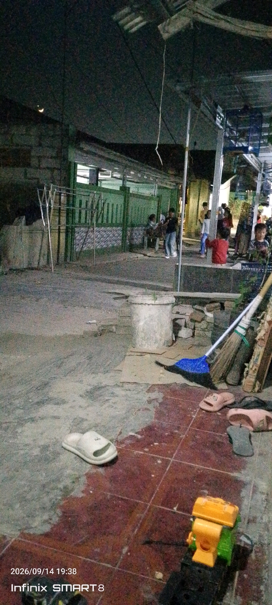 In the city of Serang, Indonesia, an outdoor scene unfolds at dusk or night, as indicated by the crescent moon visible in the upper left corner of the image. The environment appears to be a residential or communal outdoor space, possibly a yard or courtyard, with elements of both a home and public gathering area.
A green fence with vertical slats runs along the middle ground, partially obscuring a structure behind it. This structure has windows and appears to be illuminated from within, suggesting it is a building. In front of the fence, a metal clothes rack stands to the left, empty. To the right of the clothes rack, near the fence, are several people, including children and adults, some of whom are seated. One person, a child in a red shirt, is visible in the foreground-right, facing away from the camera. Other individuals are scattered throughout this area, some engaged in conversation or simply present.
The ground surface is varied. In the foreground, there is a patch of reddish-brown tile, interspersed with concrete or dirt. Scattered on the ground are several pairs of sandals, including a white pair on the left and some brown and grey pairs on the right. A blue broom with a blue dustpan is positioned near a concrete cylindrical object in the middle ground, which may be a planter or a well cover. Several bricks are stacked near this object. To the far right, a toy vehicle, predominantly yellow and green, is visible.
The overall lighting is dim, consistent with evening or nighttime. The scene suggests a relaxed atmosphere, possibly a family or community gathering after a day's activities. The timestamp at the bottom left of the image indicates "2026/09/14 19:38" and "Infinix SMART 8", identifying the device used to capture the photograph.