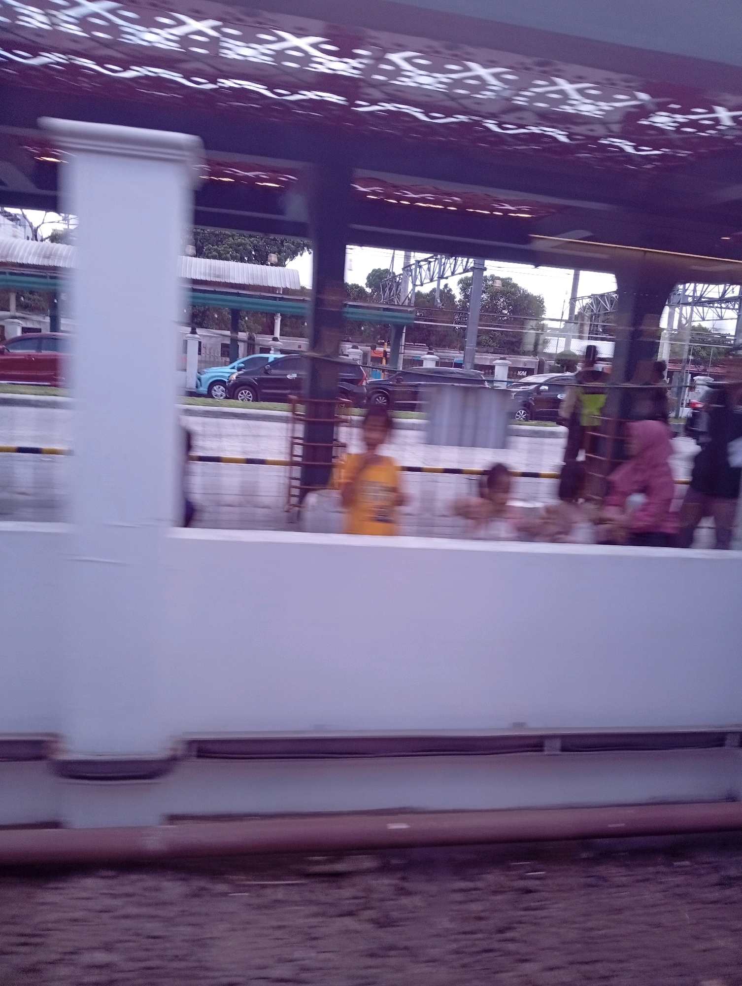 The image shows a blurry view of a street scene in Yogyakarta, Indonesia. In the foreground, a white barrier is visible. Behind it, there are people, some of whom appear to be children, standing and sitting. Some are wearing brightly colored clothing, like a child in a yellow shirt. Cars are visible in the background, including a red car, a blue car, and a black car. Overhead, a structure with patterned lighting is visible. The scene appears to be a public space, possibly near a station or transit area, given the presence of multiple vehicles and people. The lighting suggests it might be late afternoon or early evening.