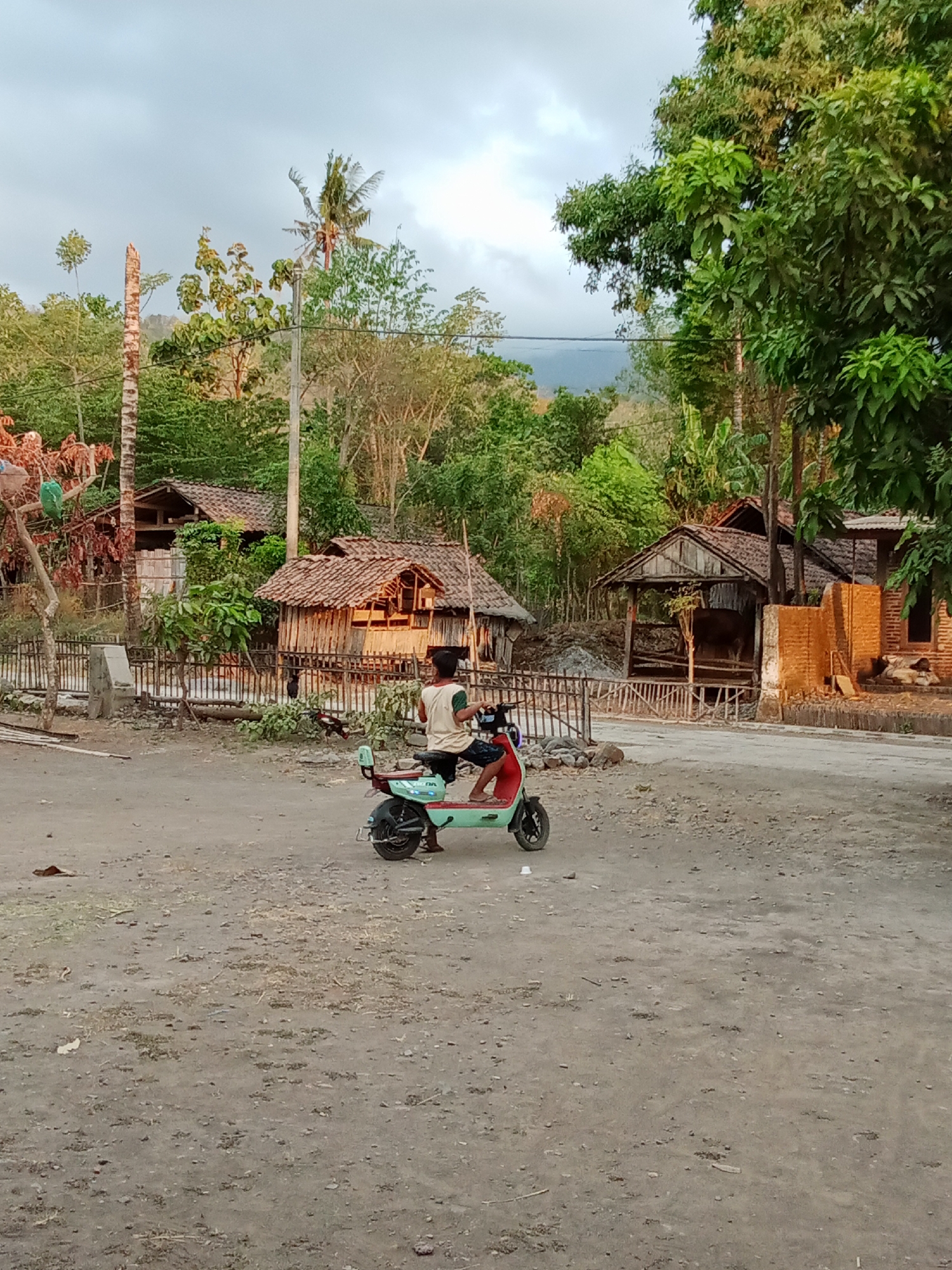 The image depicts a child riding a green and red electric scooter on a dirt road in Selowogo, Indonesia. The child, wearing a light-colored t-shirt and dark shorts, is facing away from the camera. Behind the scooter, traditional Indonesian houses with thatched roofs are visible, surrounded by lush green trees and vegetation. The sky is cloudy, suggesting overcast weather or late afternoon light. The ground is rough and dusty, with sparse grass and debris. In the background, a hill or mountain can be seen through the trees. The scene suggests a rural or semi-rural setting in Indonesia.