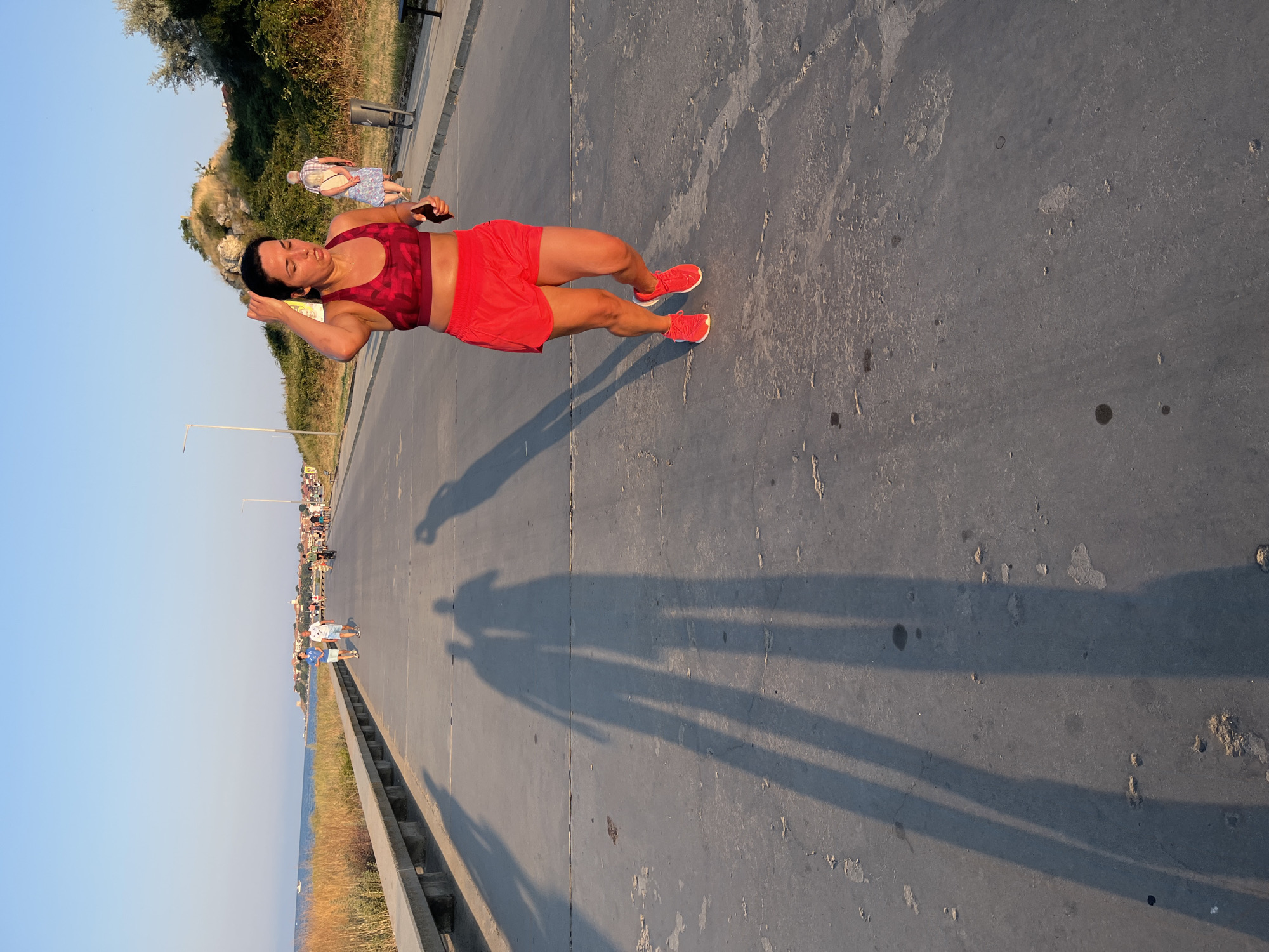 In Nesebar, Bulgaria, a woman is captured in mid-stride while jogging along a paved path beside the sea, bathed in the warm light of sunset. She wears a vibrant red sports bra and matching shorts, her hair pulled back as she touches her forehead. Her red athletic shoes are laced up, and she holds a dark object in her right hand, possibly a phone. Her long shadow stretches across the path, alongside the shadows of other people, including a couple walking in the distance and other individuals scattered further down the promenade. The background reveals a clear blue sky transitioning into a pale horizon, with the tranquil sea to the left, bordered by a strip of golden vegetation. Beyond the vegetation, a town with buildings and a church steeple is visible, suggesting a coastal urban setting. The path itself is a wide, concrete surface with visible lines and some discoloration, indicating wear and tear. The scene is peaceful, with a sense of outdoor recreation and the calm ambiance of the approaching evening.