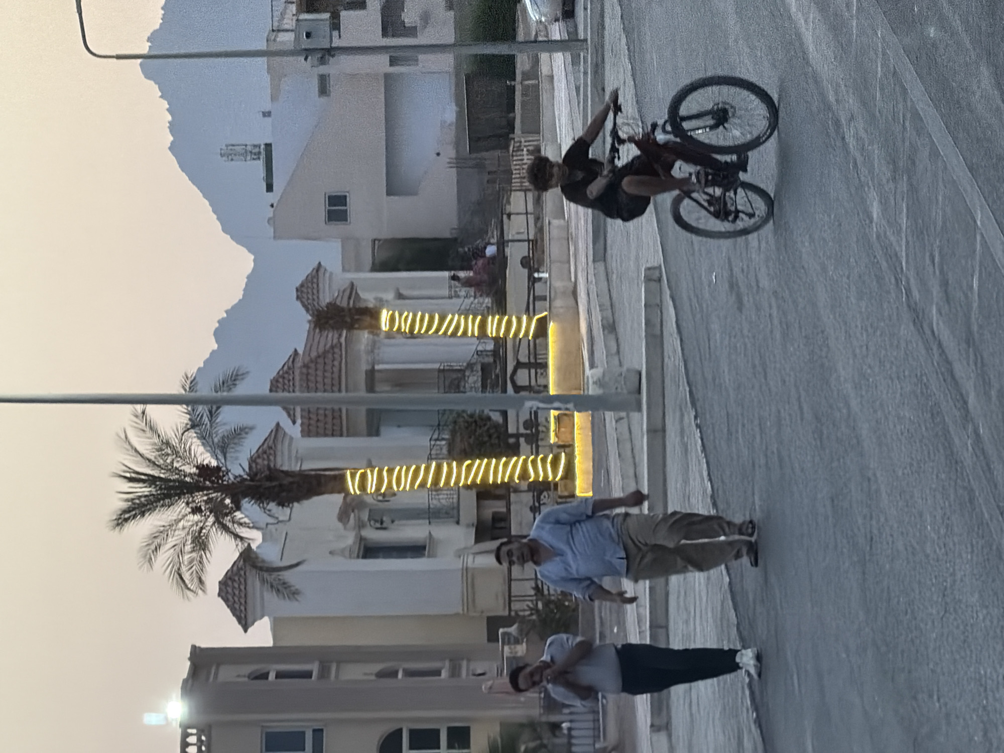 The image depicts a street scene in Sharm El Sheikh, Egypt, with a hazy sky and mountains in the background. Two men are walking on the left side of the street, dressed casually in shirts and pants. A young person is riding a bicycle on the right side of the street. Buildings with distinct architectural features, including palm trees decorated with lights, line the background, suggesting a resort or residential area. A street light is visible on the right, and the road surface is asphalt. The overall atmosphere is calm, with soft lighting indicating either dawn or dusk.