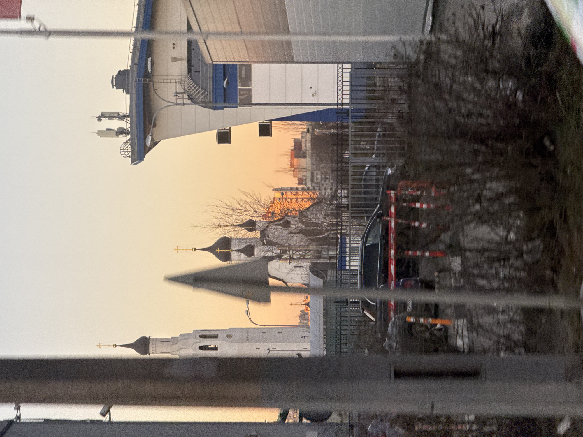 The image captures a view of Saint Petersburg, Russia, through a metallic fence or railing. The scene is oriented vertically, with the upper part of the frame showing a modern building with white and blue paneling and communication equipment on its roof. The middle ground features the silhouettes of Russian Orthodox church domes against a light orange sky, likely during sunset or sunrise. In the background, city buildings are visible, reflecting the ambient light. The lower part of the frame is dominated by the fence, which obscures the immediate foreground. A dark vehicle and construction barriers are also partially visible behind the fence. The overall impression is a juxtaposition of modern and historical elements within the urban landscape.