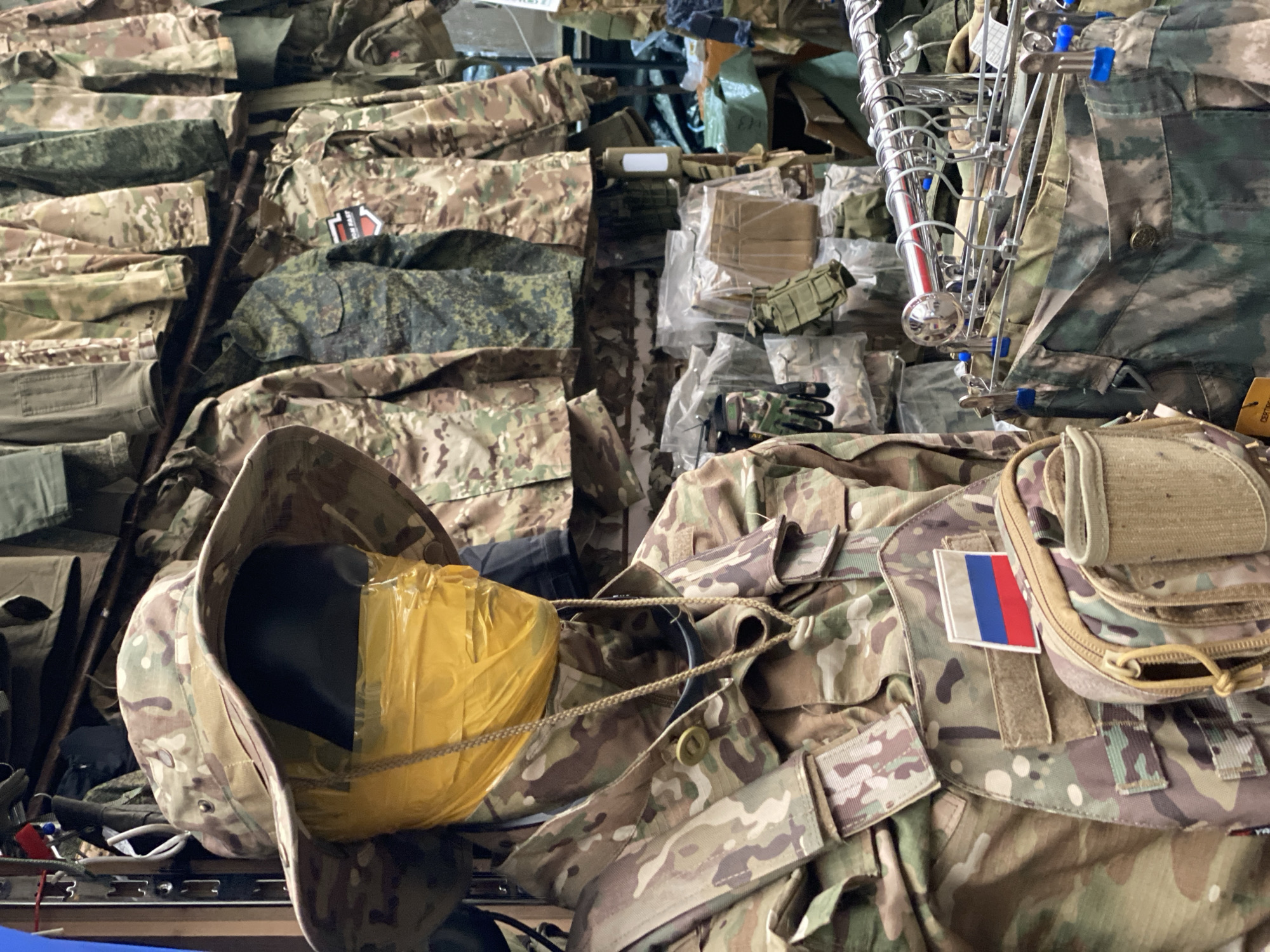 The image shows a store display of military-style clothing in Moscow, Russia. A mannequin in the foreground is dressed in a camouflage uniform, complete with a wide-brimmed hat, a vest with a Russian flag patch, and pouches. The mannequin's face is obscured by yellow tape. Behind the mannequin, racks of similar camouflage clothing, including jackets and pants, are displayed. Various bags and tactical gear are also visible, some in plastic packaging. The overall impression is of a retail environment selling military apparel.