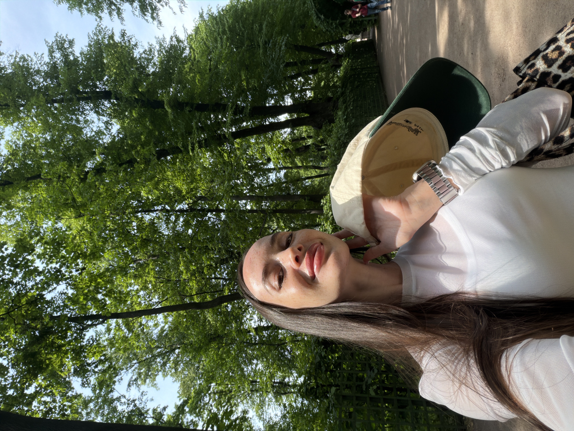 A young woman is taking a selfie in a park in Saint Petersburg, Russia, on a sunny day. She has long, dark brown hair and is wearing a white, sheer top. She is holding a beige baseball cap with a green brim in her left hand and resting her chin on her right hand. She has a silver watch on her left wrist. She is smiling, and her face is lit by the sun.
The background is a lush green park with tall trees and a pathway. In the distance, two men are walking on the pathway. The woman is standing in the foreground, with the park and trees behind her. The overall atmosphere is bright and cheerful.