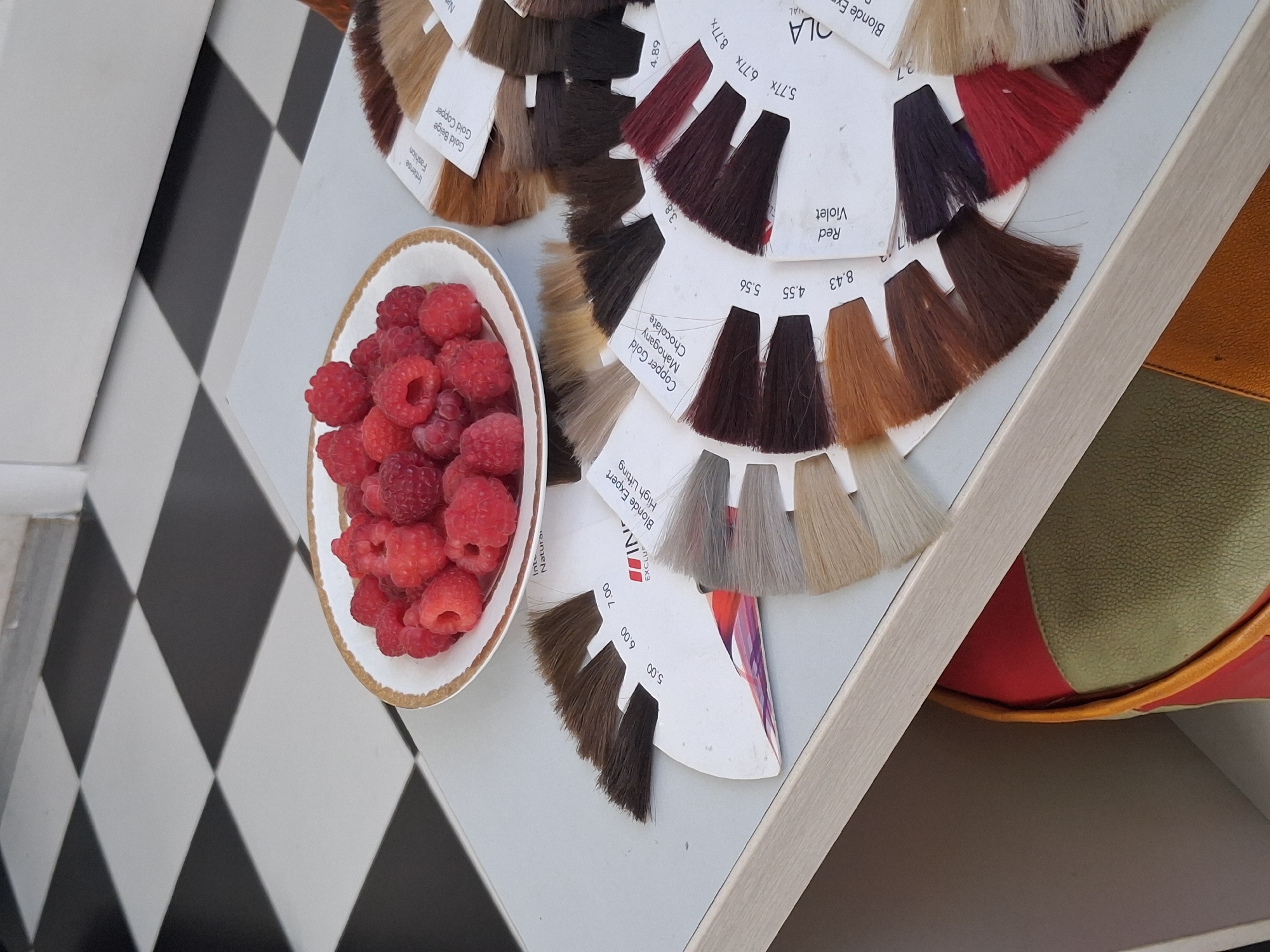 In the city of Tuzdybastau, Kazakhstan, a close-up, top-down shot reveals a white table surface adjacent to a black and white checkered tiled floor. On the table sits a white porcelain bowl filled with fresh raspberries. To the right of the bowl are several fan-shaped color swatches, likely for hair dye, arranged in a semi-circular pattern. Some swatches display hair strands in various shades, while others have printed labels indicating colors and numbers such as "Intense Fashion," "Gold Beige Gold Copper," "Natural," "Copper Cold Mahogany Chocolate," "Red Violet," and numerical codes like "5.00," "6.00," "7.00," "3.8," "4.89," "5.56," "8.43," "4.55," and "8.77x." Underneath the table, a glimpse of a red and green object, possibly a bag or cushion, is visible within a shelf or recess.