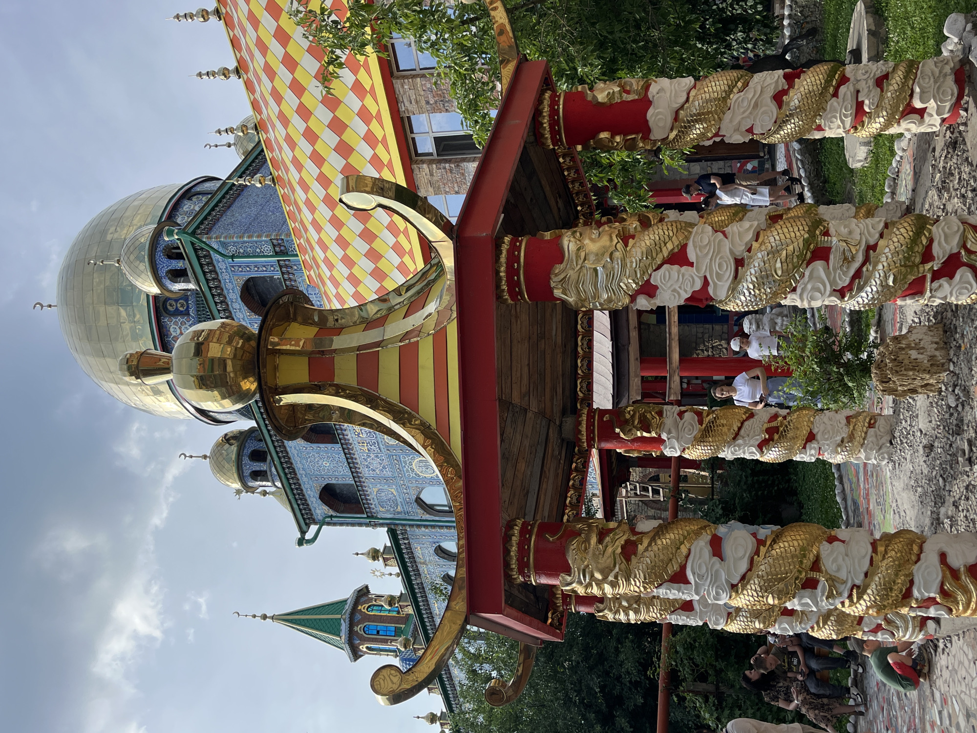 This image captures a view of the Temple of All Religions in Kazan, Russia. In the foreground, a red and gold gazebo with dragon-shaped pillars stands prominently. The gazebo's pillars are adorned with golden dragons wrapped around red and white sections, resembling clouds. Beyond the gazebo, the multi-religious complex is visible, featuring a large golden dome, a green spire, and a roof covered in a checkered pattern of yellow, red, and orange tiles. The building's facade is decorated with intricate blue and white tilework. Several people are present in the scene, including a woman in a white top and blue skirt standing near the gazebo, a group of people sitting on a bench to the left, and a child crouching on the ground in the lower left corner. The environment is lush with green trees and foliage surrounding the structures. The sky above is partly cloudy.