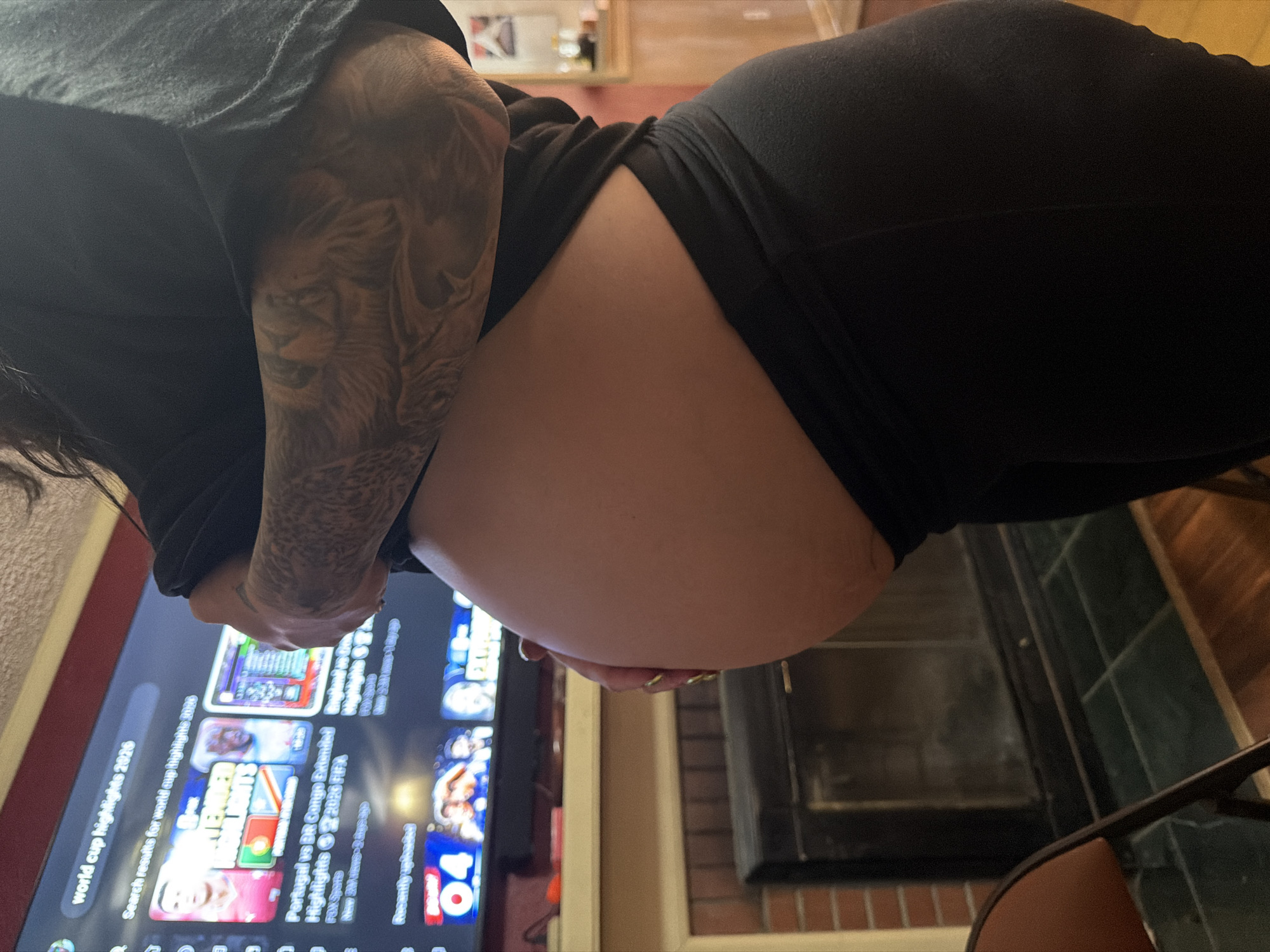 A person with a visible pregnant belly, wearing black clothing, is standing in front of a television screen displaying search results for "world cup highlights 2026." The television is situated above a fireplace with a dark insert. To the left of the fireplace, a chair is partially visible. The scene appears to be indoors, likely in a residential setting. The location is the city of Virginia Beach, United States of America.