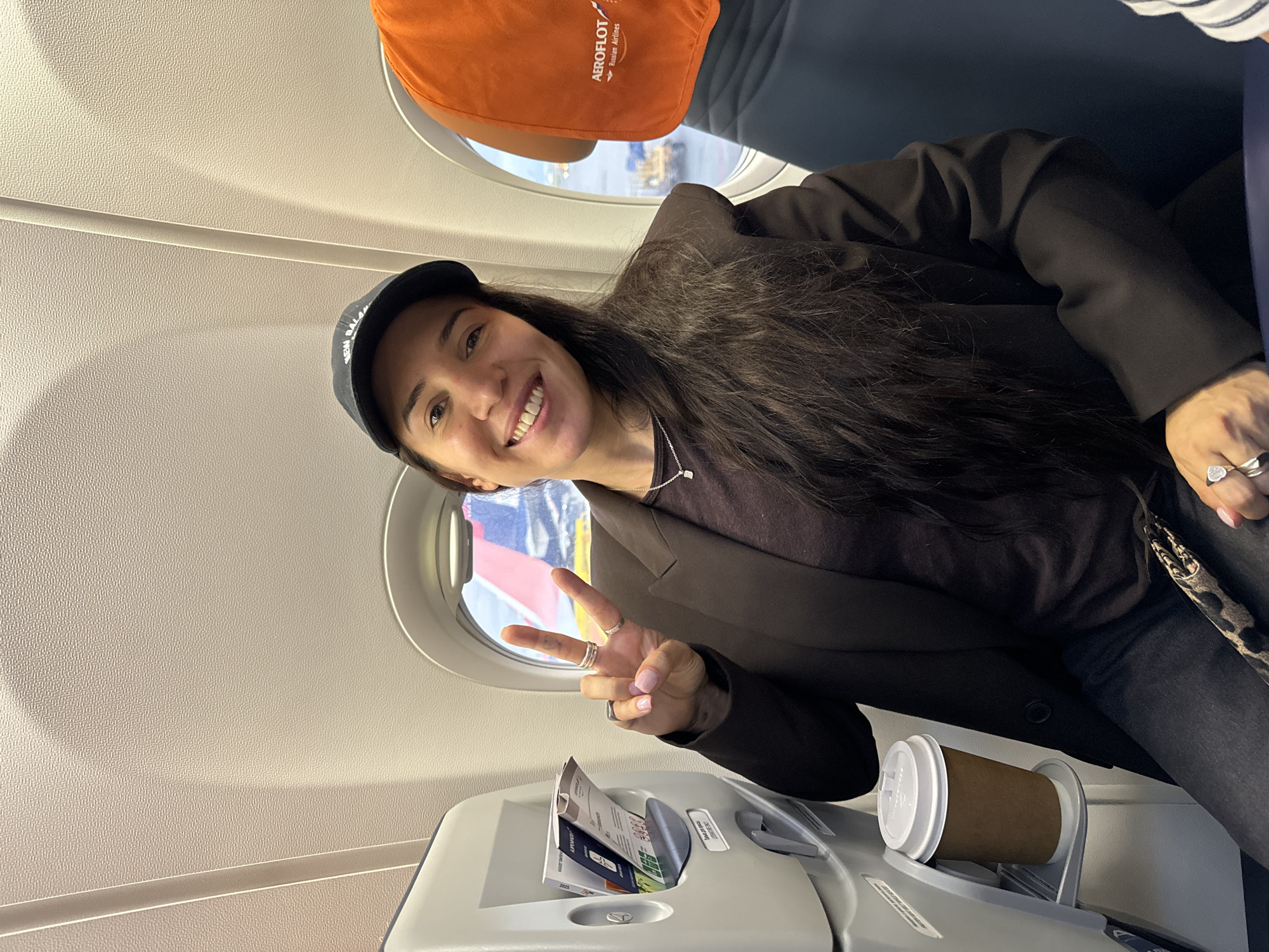 A woman smiles and flashes a peace sign while seated on an Aeroflot Russian Airlines airplane. She wears a black baseball cap, black jacket, and a dark top. The airline seat has an orange headrest cover with the Aeroflot logo. In the seat in front of her is a cup of coffee and a tray containing airline pamphlets. Through the window, the cityscape of Khimki, Russia, is visible.