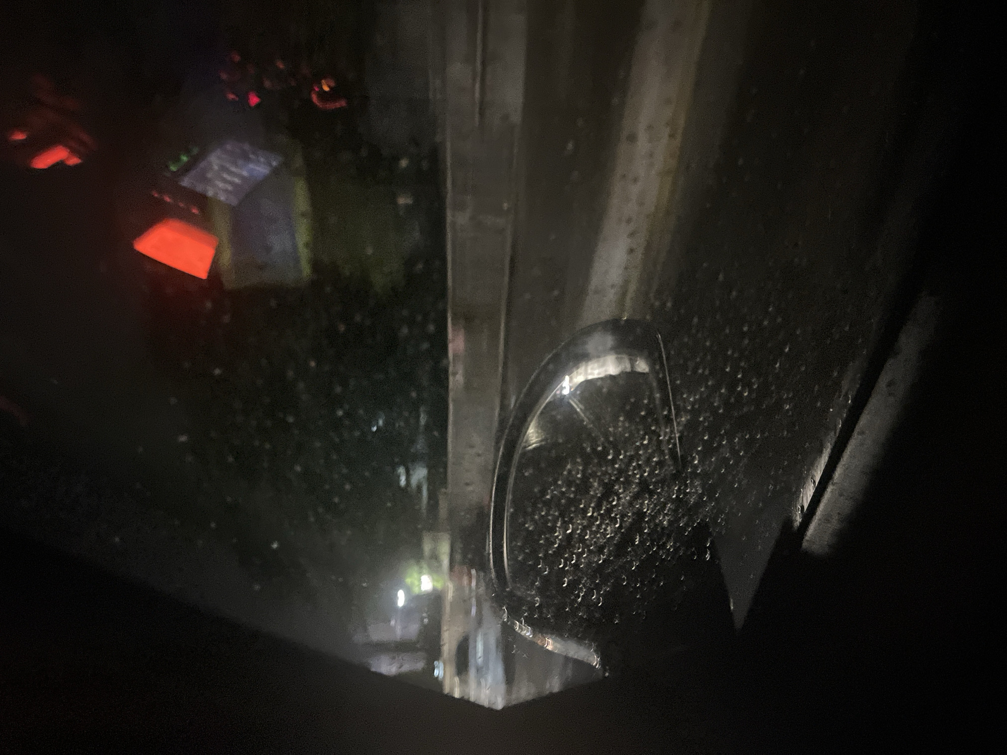 The image depicts a dark, rainy night scene in an urban environment, viewed through a wet, possibly glass, surface covered in numerous water droplets. The perspective is from a slightly elevated position, looking down into a dimly lit area.
In the mid-ground, a prominent metallic, curved structure, possibly part of a streetlamp or architectural element, is visible on the right side, reflecting ambient light. Various straight, dark structural elements, resembling parts of a building, a bridge, or an elevated platform, run vertically and horizontally across the upper and central portions of the frame.
The background is largely obscured by darkness and atmospheric conditions. However, several artificial light sources punctuate the gloom. To the top-left, blurred red and orange glows suggest traffic lights or illuminated signs. Below these, a rectangular, illuminated object with faint green and white elements appears to be a digital display, possibly from an ATM or an information screen, but no specific text is legible. Further to the bottom-left, indistinct bright spots indicate distant streetlights or building lights. No people or discernible activities are present. The scene conveys a quiet, reflective mood typical of a rainy night in a city like Blagoevgrad, Bulgaria.