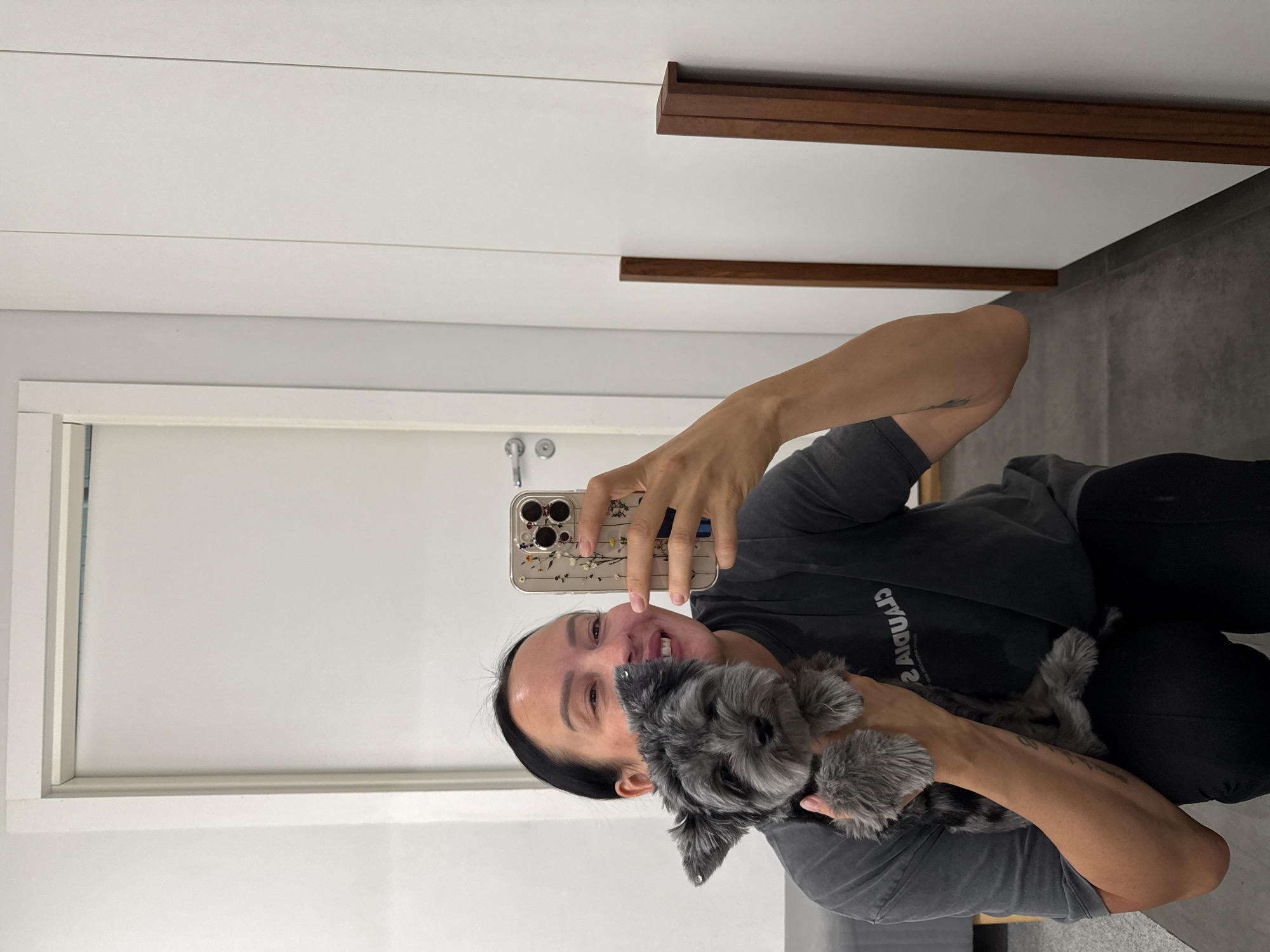 A woman with dark hair and light skin is holding a small, grey dog. She is smiling and taking a selfie with her phone. She is wearing a grey t-shirt and black pants. The dog appears to be a terrier mix with a scruffy coat. They are in a room with white walls and a white door. There are also two wooden vertical beams on the wall. The floor is grey. The image does not contain any information about the location being the city of Chaikovsky, Russia.