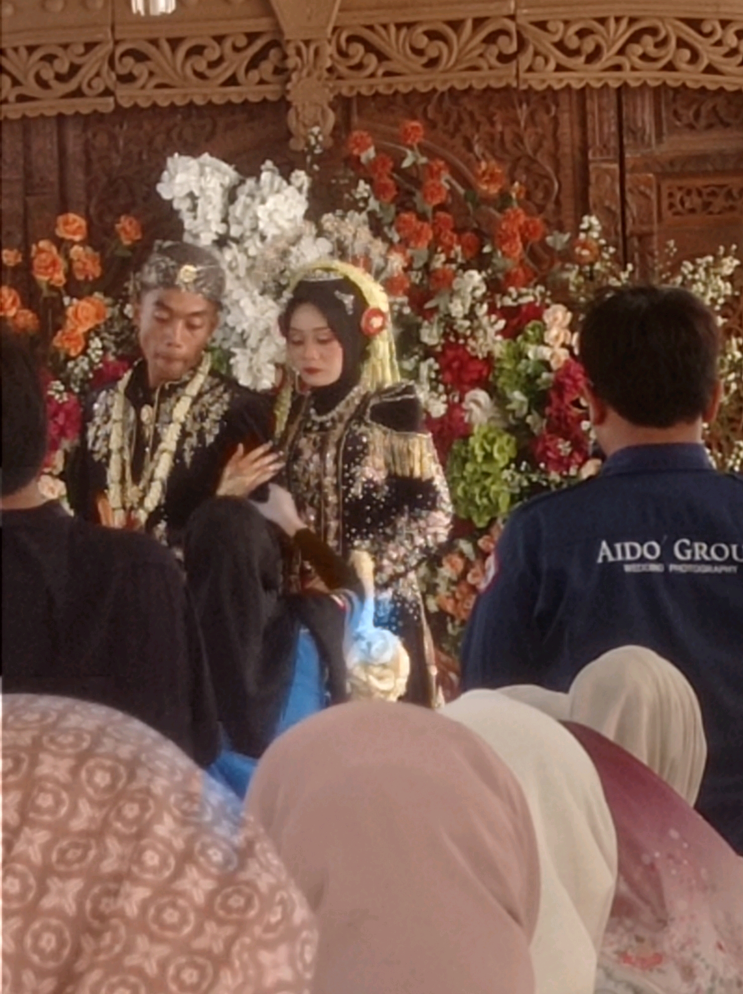 A wedding ceremony is taking place in Ngemplak, Indonesia. The bride and groom are seated in front of a floral backdrop and intricately carved wooden decorations. The groom is wearing a traditional black outfit with gold embellishments and a lei of white flowers. The bride is dressed in a black and gold patterned wedding dress with a yellow headscarf and veil. A photographer in a blue uniform with the text "Aido Group Wedding Photography" on the back is also present. Several guests, mostly women wearing headscarves, are seated in the foreground, partially obscuring the bride and groom. The overall atmosphere appears to be solemn and traditional.