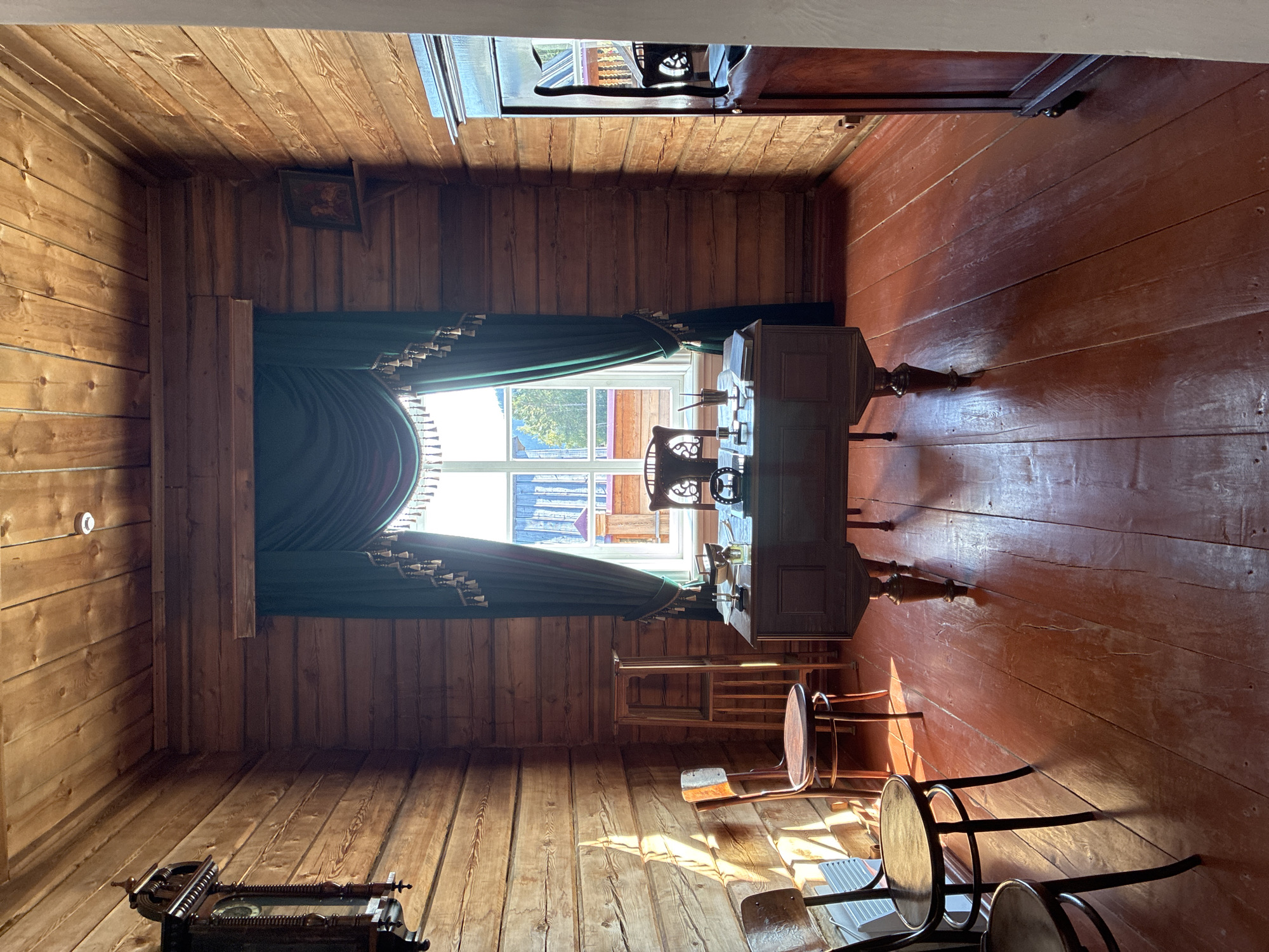 This image depicts a room in a historical wooden building, likely a museum, located in the Taltsy (Irkutsk) Museum of Wooden Architecture in Russia. The room is furnished as a study or office, with wooden plank walls and ceiling. Dominating the center of the room is a dark wooden desk with an ornate design, placed in front of a large window draped with green curtains. On the desk are several objects, including a pen holder with pens, a book, and what appears to be a small glass bottle. A matching wooden chair is positioned behind the desk.
To the left of the desk, bathed in sunlight streaming from the window, are two wooden chairs, one positioned behind the other. A tall, narrow wooden stand with shelves is visible between the chairs and the desk. A large, dark grandfather clock hangs on the left wall, its face partially obscured by shadow.
On the right side of the room, a tall, dark wooden cabinet or wardrobe stands against the wall. A small framed picture is mounted on the wooden wall above a shelf to the right of the window. The floor is made of wide wooden planks painted red, reflecting the sunlight and the warm tones of the wood. The overall impression is of a preserved historical interior, offering a glimpse into past domestic or professional life.