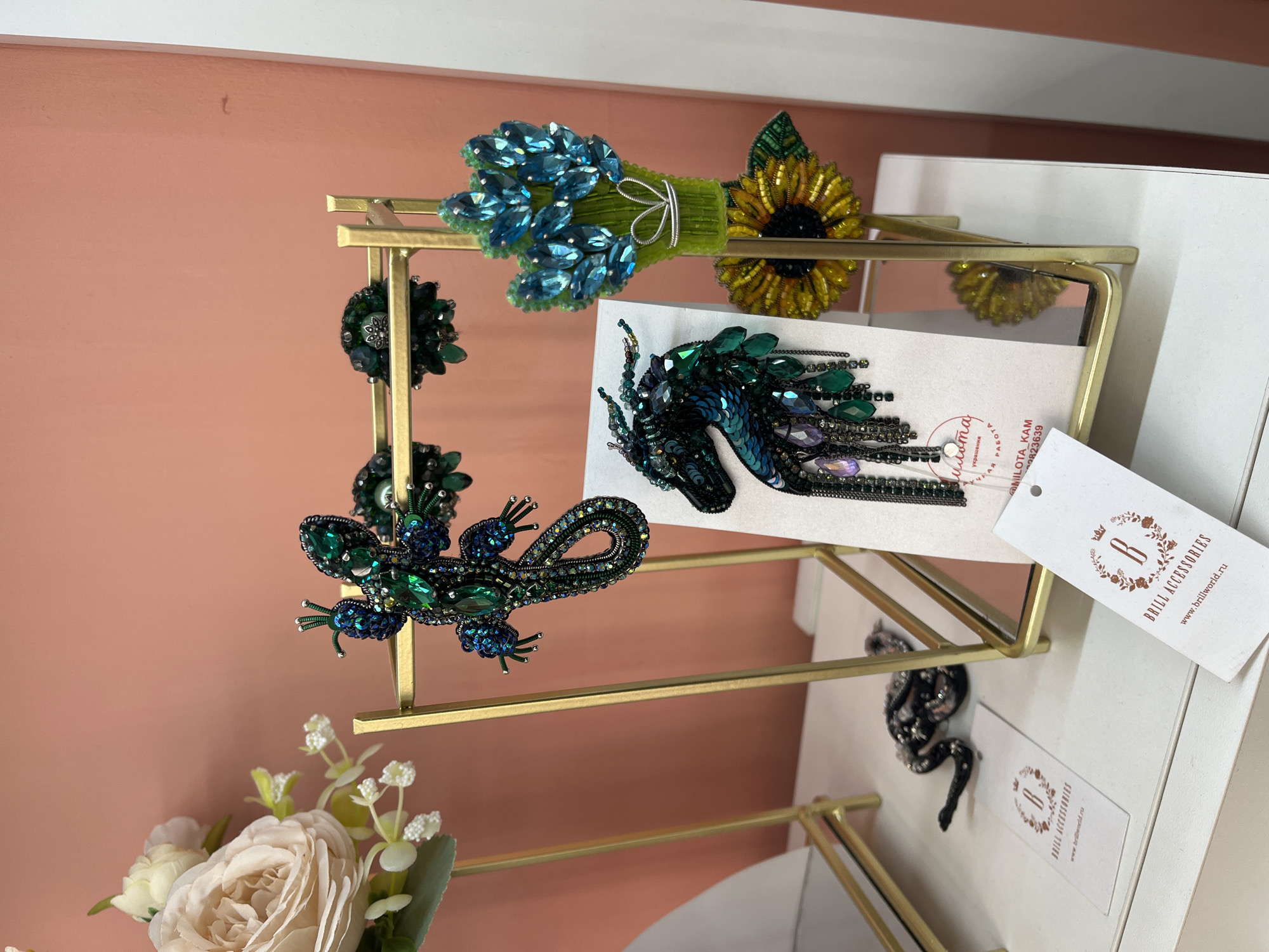 An artistic display of beaded and jeweled brooches is presented on a gold metal stand. A bouquet of blue faceted gems and a sunflower brooch are prominently featured on the right side of the stand. To the left, a detailed green lizard brooch hangs, with another smaller green brooch above it. The brooches are designed with intricate beadwork and sparkling stones, evoking a sense of luxury and craftsmanship. The background is a soft, peachy-pink wall, suggesting an indoor setting like a boutique or a craft fair. In the foreground, a white surface with several tags bearing the logo "Brill Accessories" is visible, indicating the brand or seller. A partially visible black sequined snake-like object lies on the surface, possibly another piece of jewelry or a decorative item. The overall impression is one of a curated collection of handmade jewelry. The specific location within the city of Svetly, Russia, is not identifiable from the visual information provided.
