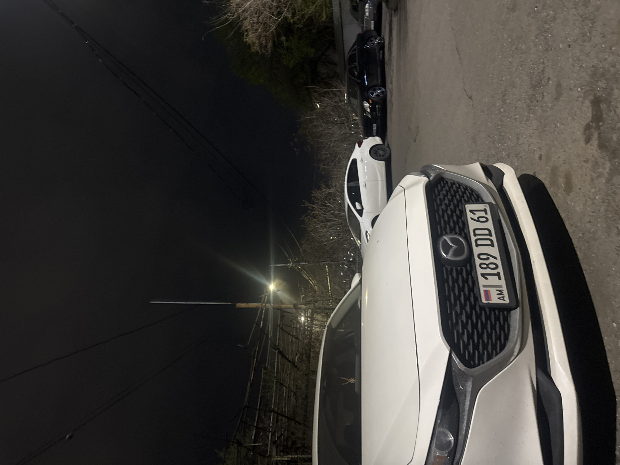 The image depicts a nighttime scene in Yerevan, Armenia. Focus is on the front of a white Mazda CX-5, with Armenian license plate "189 DD 61" prominently displayed. Parked adjacent to the Mazda is another white car, partially visible. Further down the street, two dark-colored vehicles are parked. Overhead power lines extend from the lower-left corner toward a streetlight illuminating the scene. Street and sidewalk are asphalt. Bare trees and vegetation line the street. The sky is dark.