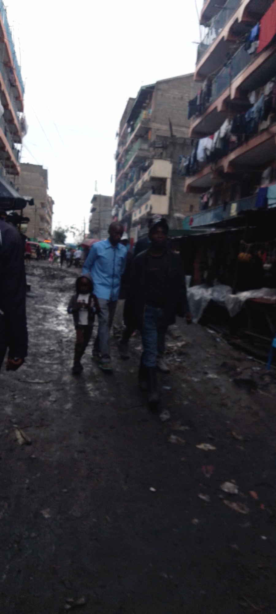 The image depicts a muddy street scene in what appears to be an informal settlement in Nairobi, Kenya. Several individuals are walking along the street, including a child. Multi-story buildings flank both sides of the street. Some have laundry hanging from the balconies. Market stalls or informal businesses are visible along the right side of the street. The ground is muddy and uneven. The sky is overcast.