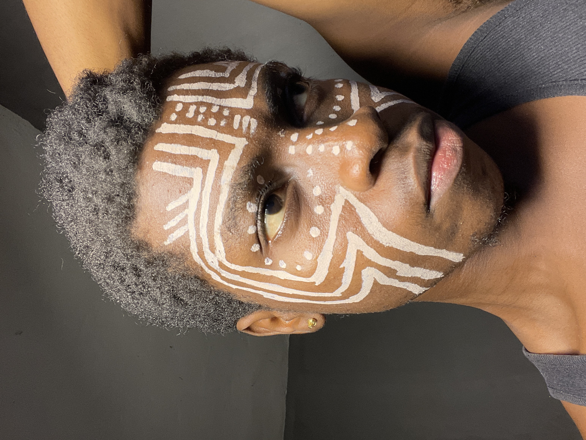 A close-up, portrait-style photograph depicts a person with dark skin and a shaved, gray-tinged hairstyle, lying on their back against a dark gray surface, presumably a wall or floor. The person's face is adorned with white painted designs resembling tribal or cultural markings, featuring linear patterns and dots. A gold earring is visible in the person's ear. The person wears a gray garment, and part of their arm is positioned above their head. There is no evidence to suggest it was taken in Nkpor, Nigeria.