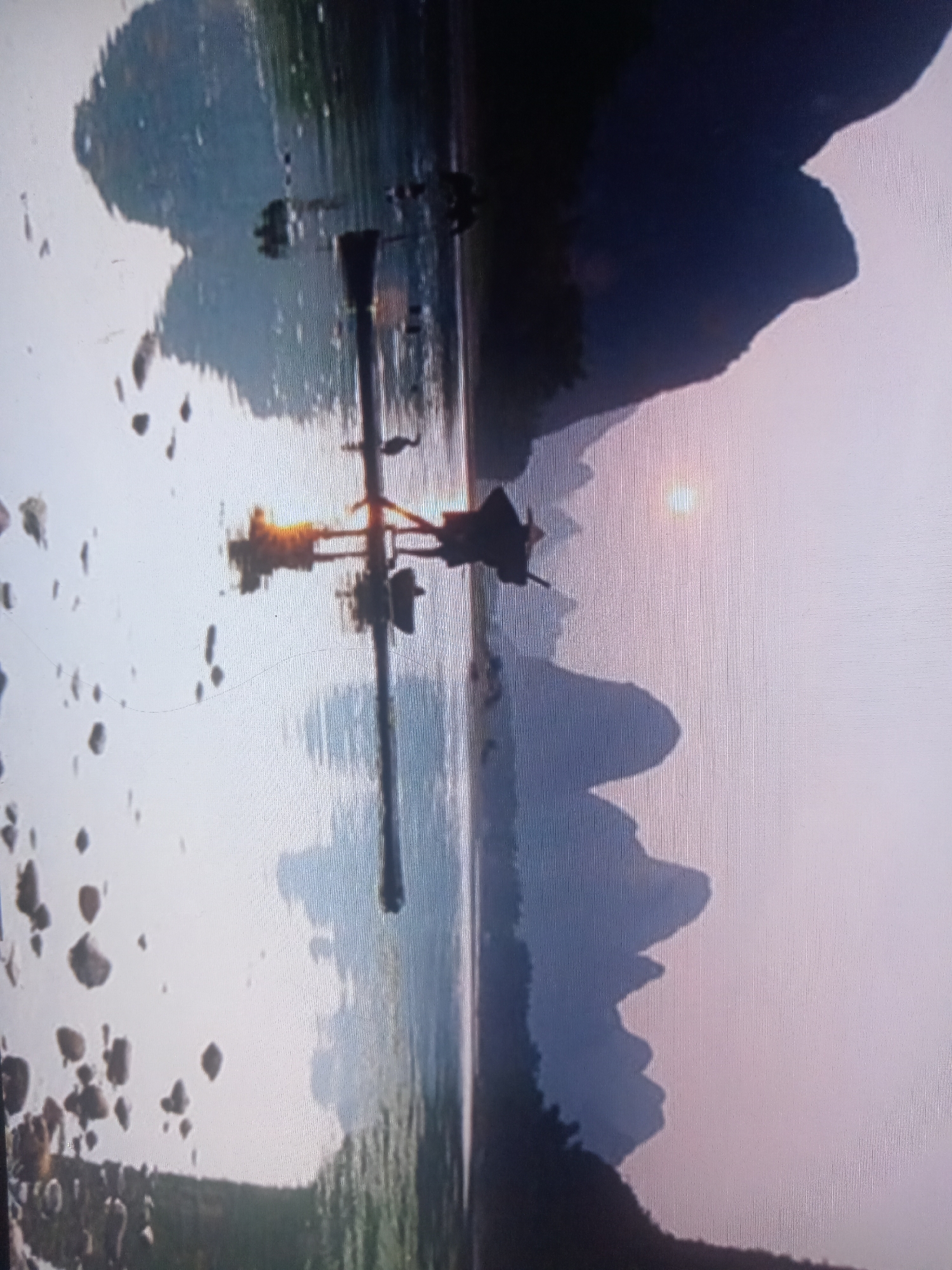 The image depicts a blurry, vertical landscape with a body of water reflecting mountains and a hazy sun. A structure, possibly a boat or a pier, with a tall, thin pole and multiple crossbars, is visible in the center of the frame. The pole is topped with a dark, angular object. The water is disturbed, with ripples and reflections of nearby rocky outcrops on the left. The background features silhouetted mountains against a pale sky. The overall impression is a serene, natural scene with a man-made element interrupting the landscape. There are no discernible people or explicit actions taking place.