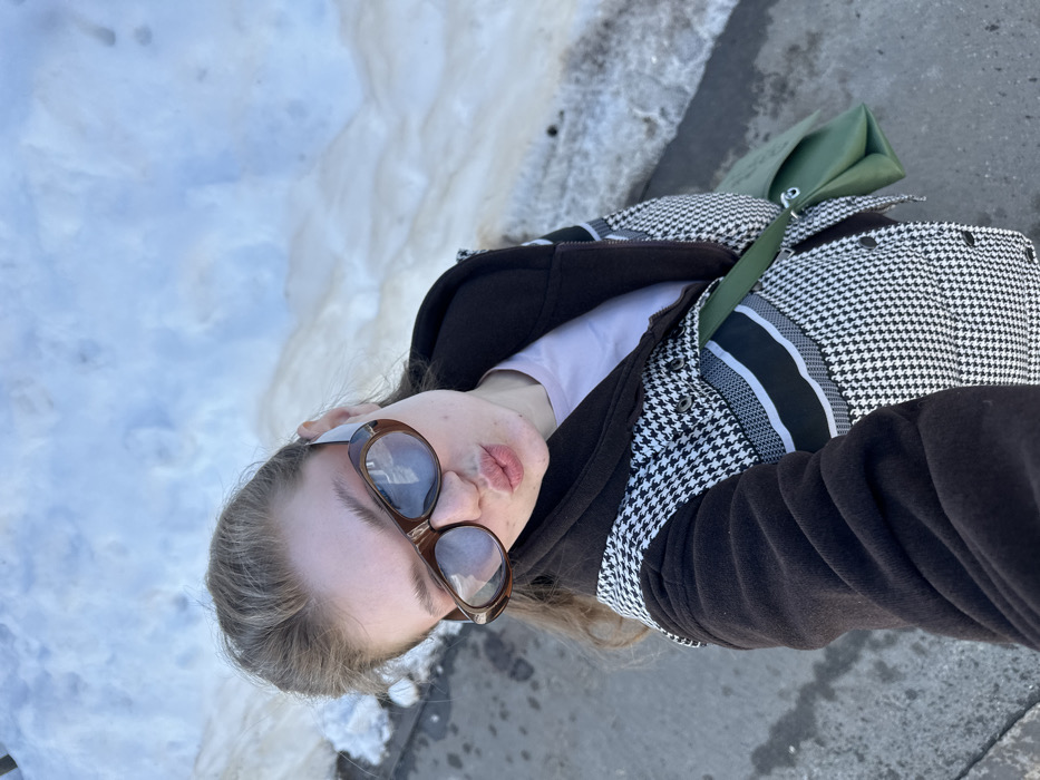 A forensic description of the scene in Новое Шигалеево, Russia:
An outdoor, high-angle selfie captures a female individual from the chest up, posing with pursed lips. The subject wears oversized brown-framed sunglasses with reflective lenses, partially obscuring her eyes. Her light brown hair is pulled back. She is dressed in a dark brown jacket featuring a prominent black and white houndstooth pattern across the torso, with solid dark brown sleeves and collar, over a light pink t-shirt. A green shoulder bag is worn across her body, visible on her right side. The background consists of a large accumulation of snow, exhibiting patches of melting and dirt, indicating cold weather conditions. A strip of dark grey pavement or asphalt is visible along the lower left edge of the frame. The scene is brightly lit by natural sunlight.