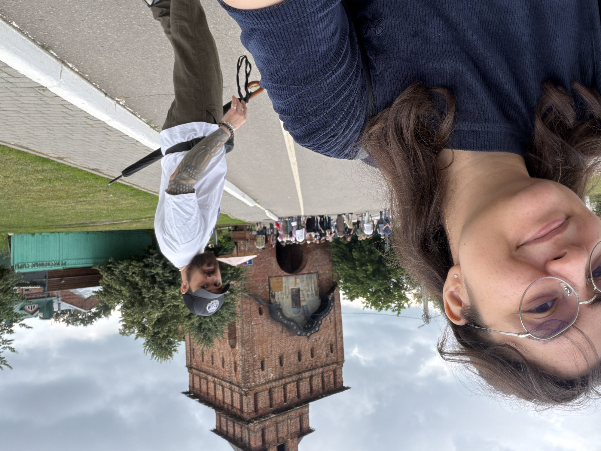 In Kolomna, Russia, a woman with glasses and dark hair smiles at the camera, her shoulder and chest filling the foreground. Behind her, a man with a beard wears a white t-shirt, a black baseball cap, and dark pants, holding a closed umbrella. They are on a paved path in front of a large, historic brick tower, likely a part of the Kolomna Kremlin. A crowd of people walks along the path toward the tower, and a green fence with text is visible in the background, alongside trees and buildings. The sky is overcast.