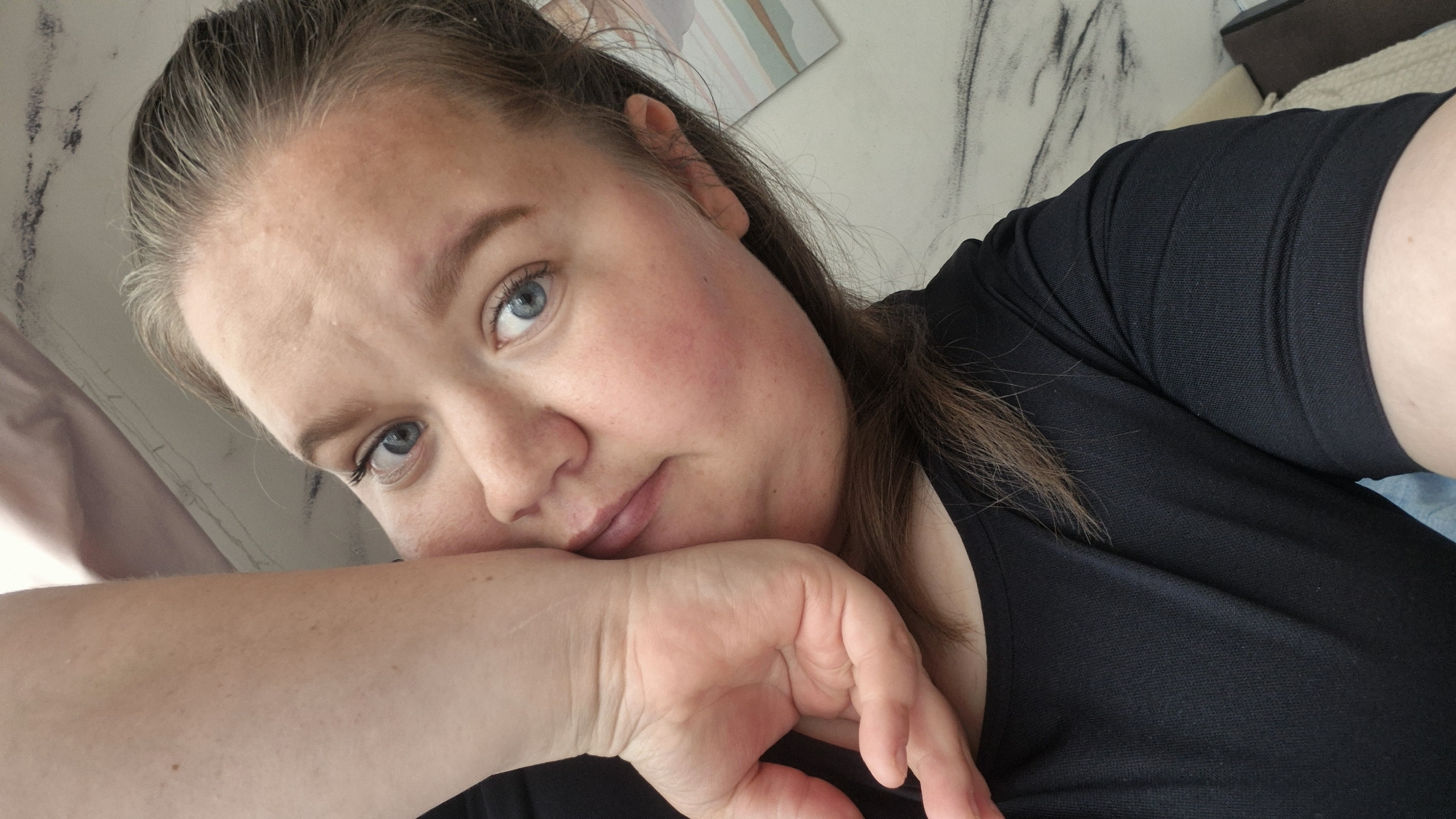 This is a selfie of a young woman with light skin, blue eyes, and light brown hair pulled back, likely taken indoors. She is resting her head on her left arm and hand. Her expression is neutral. She is wearing a black t-shirt. The background appears to be a bedroom with a light-colored wall and some abstract art or wallpaper. The image does not contain any information that could help determine its location in the city of Chaikovsky, Russia.