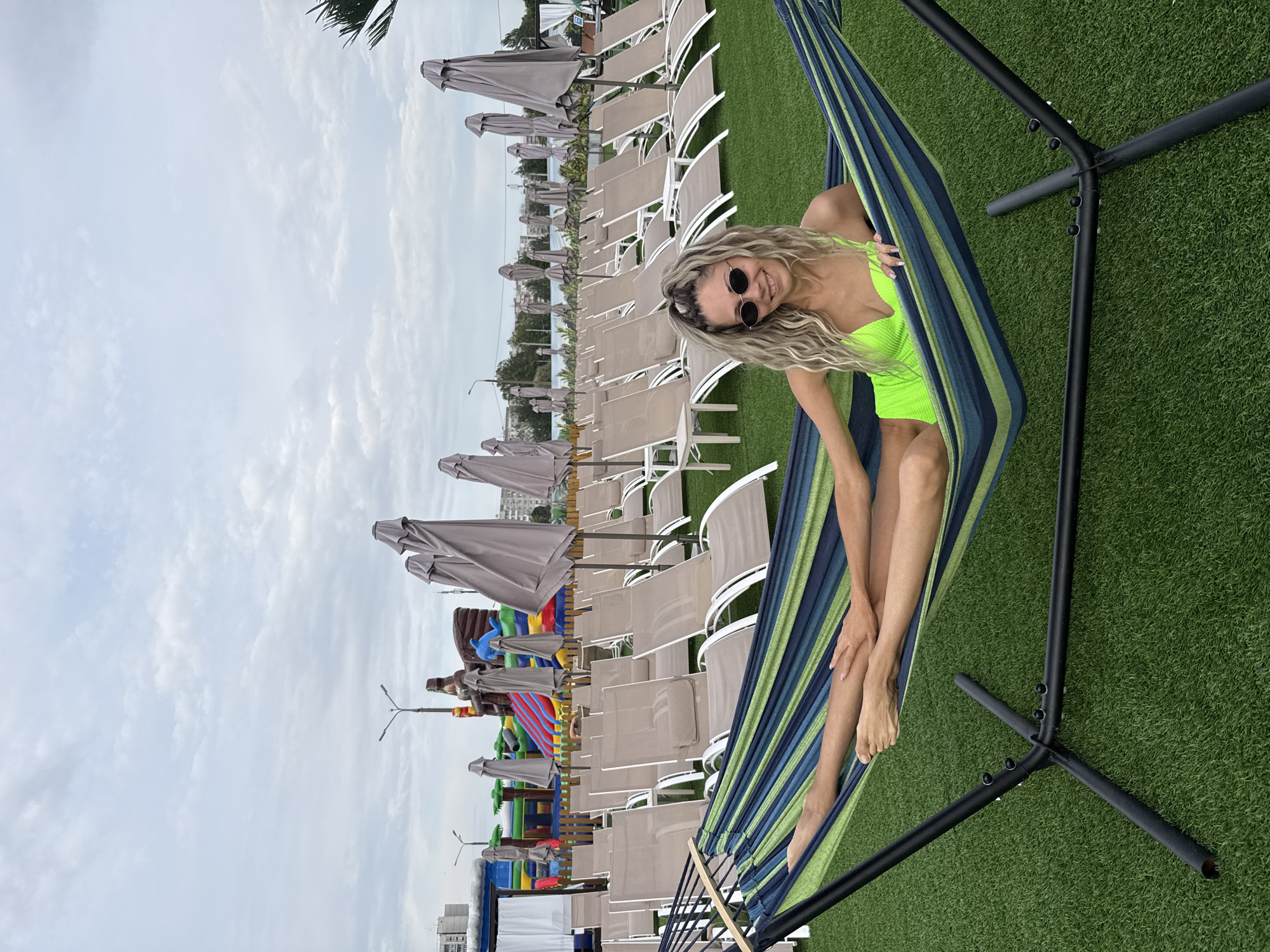 A woman with blonde, curly hair, wearing sunglasses and a neon green swimsuit, is relaxing in a striped hammock at an outdoor venue in Rusanovo, Russia. The hammock is suspended by a black metal frame and rests on artificial green turf. Behind her, rows of empty lounge chairs are neatly arranged on the turf, with closed beach umbrellas positioned between them. In the background, a colorful inflatable playground with slides is visible, along with buildings and trees. The sky is overcast.