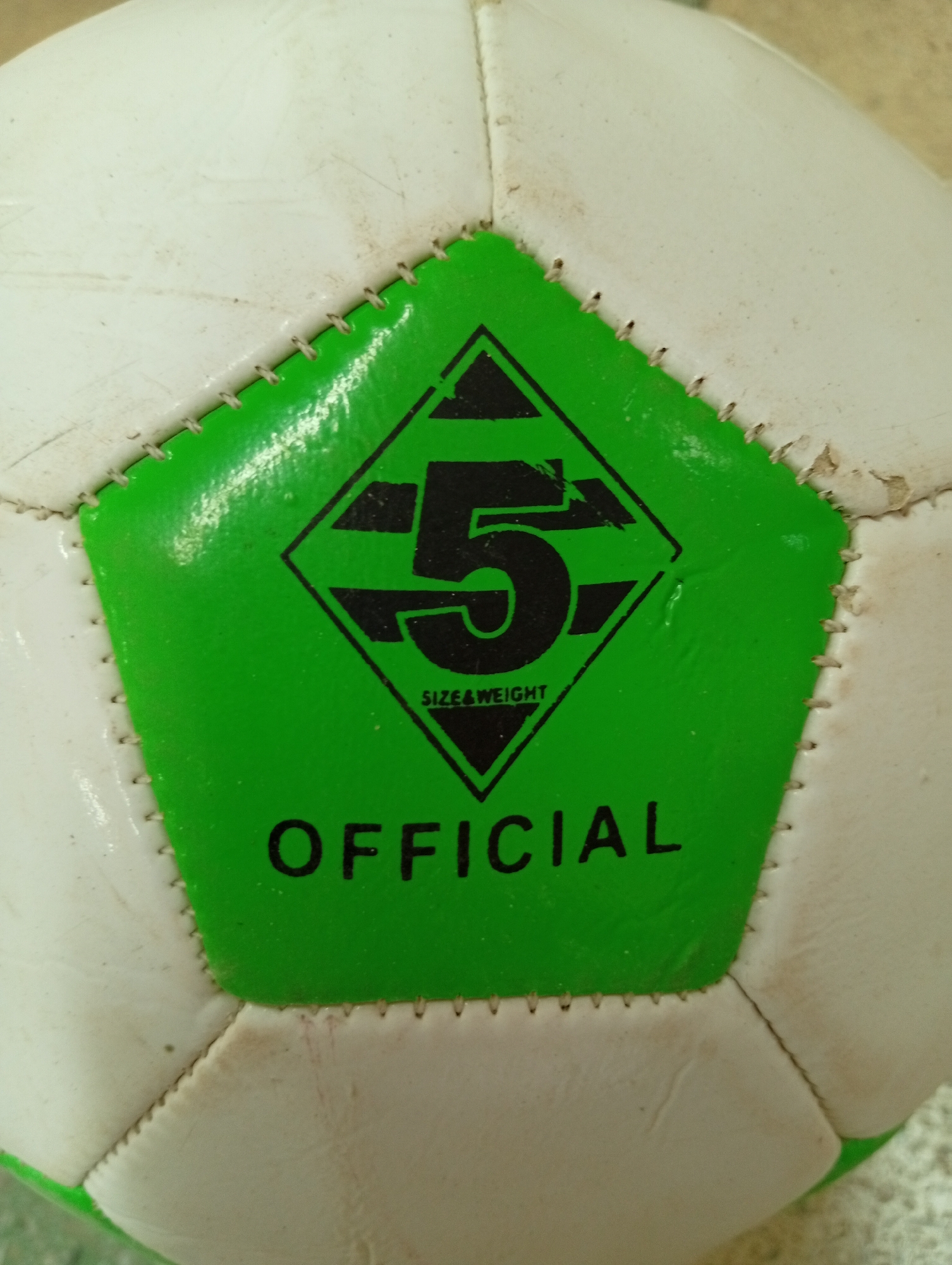 The image shows a close-up view of a white and green soccer ball. The ball is stitched with white thread and has a pentagonal green panel in the center. On this panel, there is a black diamond shape with a black number "5" inside. Below the number "5," the words "SIZE & WEIGHT" are visible in smaller black text. Below the diamond shape, the word "OFFICIAL" is written in large black letters. The background is out of focus and appears to be a textured surface, possibly a table or floor. The image does not provide any information about a specific location in Крыўлянскі сельскі Савет, Belarus.
