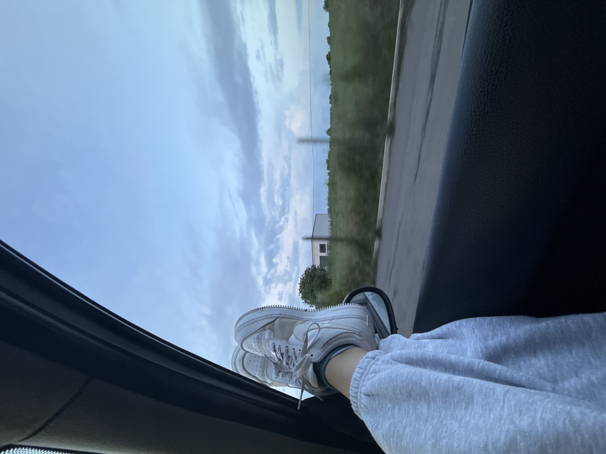 From the vantage point inside a car, a pair of white sneakers rest on the open car window, with a leg clad in grey sweatpants visible. Outside the window, a passing landscape features a grassy field, road, a white building, trees, and a cloudy sky. It is difficult to assess the location as Burgas, Bulgaria, without additional information.